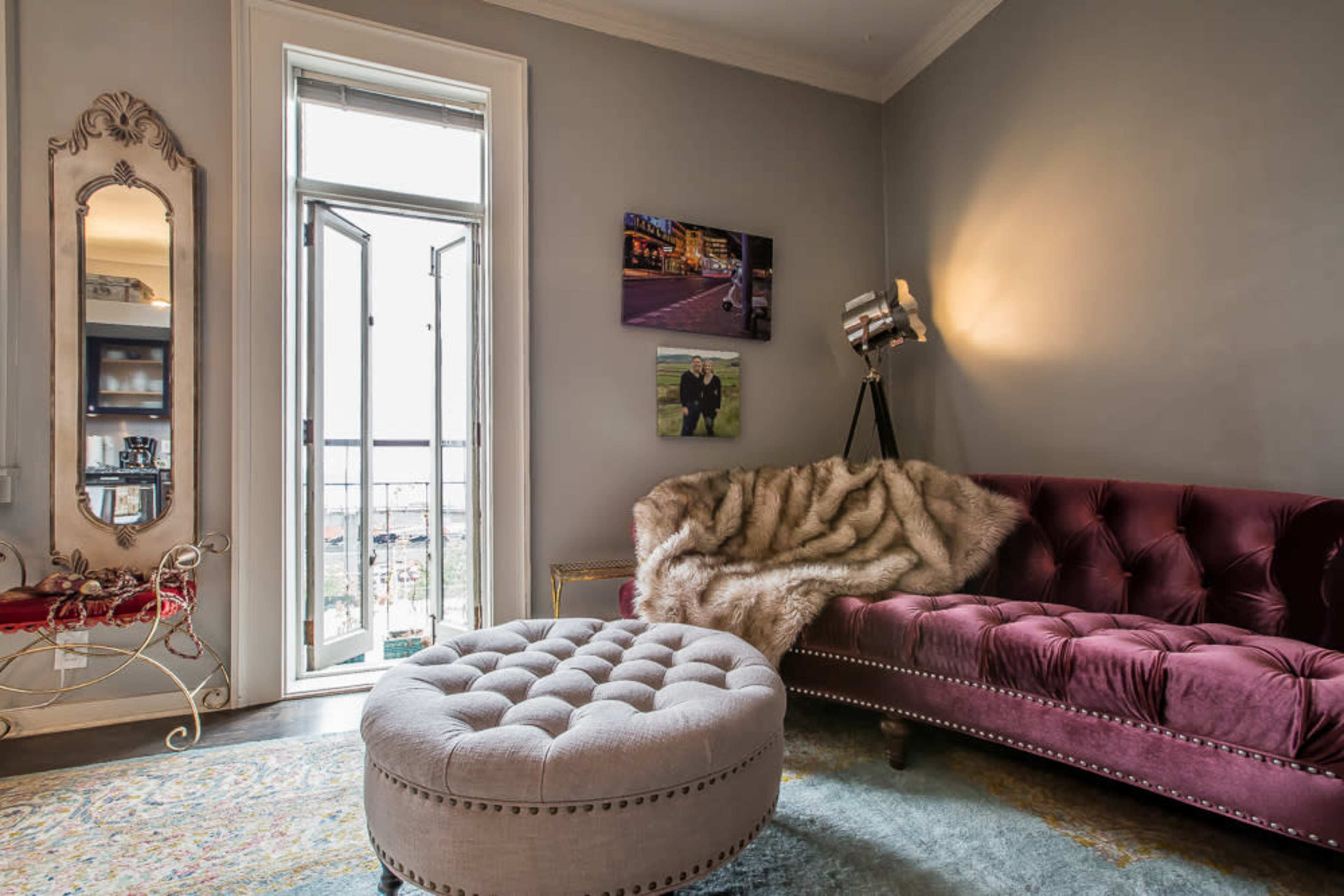 The image shows a cozy living room with a purple tufted sofa, a circular upholstered ottoman, and a balcony door allowing natural light to enter.