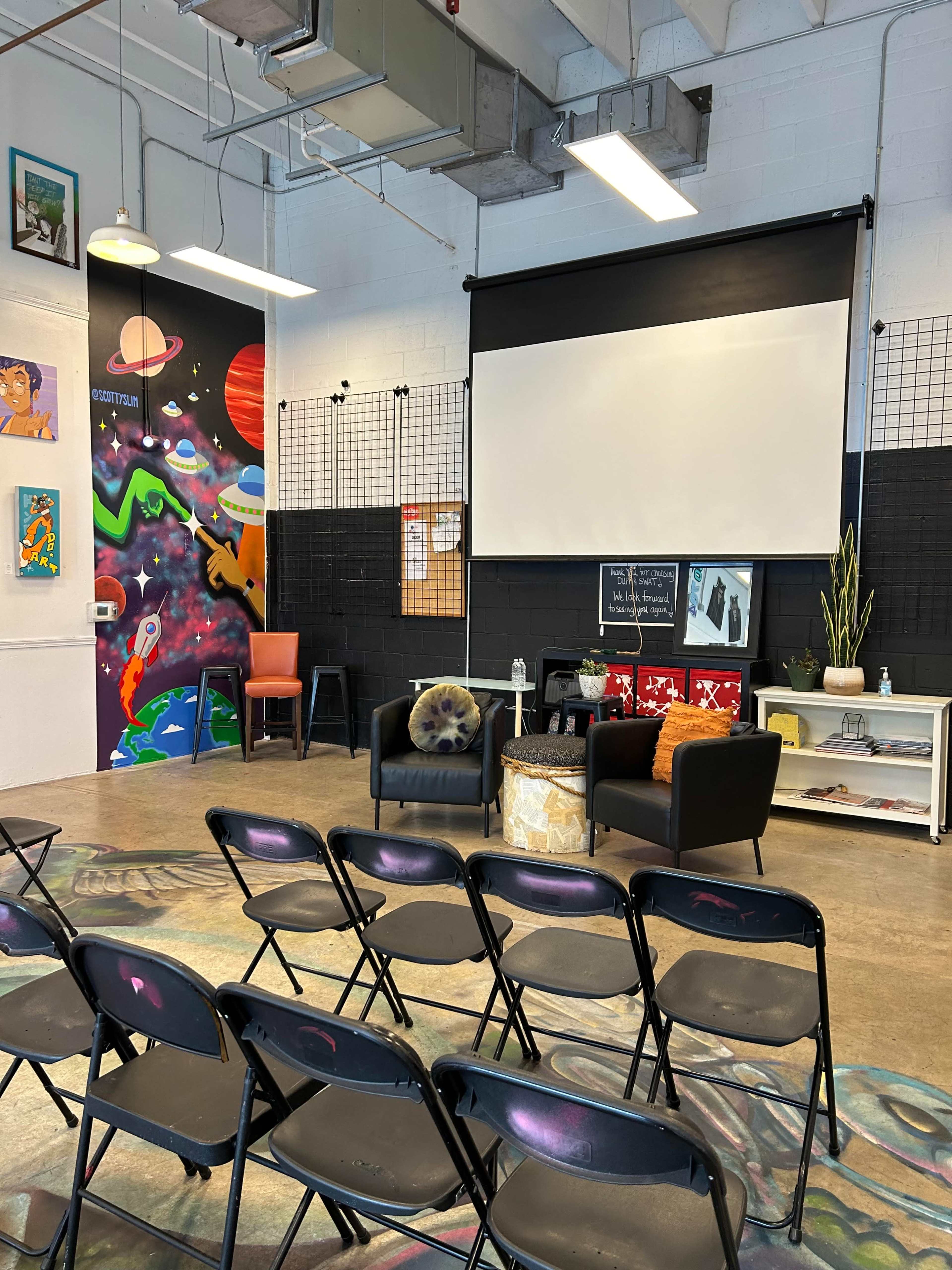 The image shows a brightly lit event space with rows of black chairs facing a white projection screen, colorful wall art, and a few pieces of furniture, including chairs and a small table.