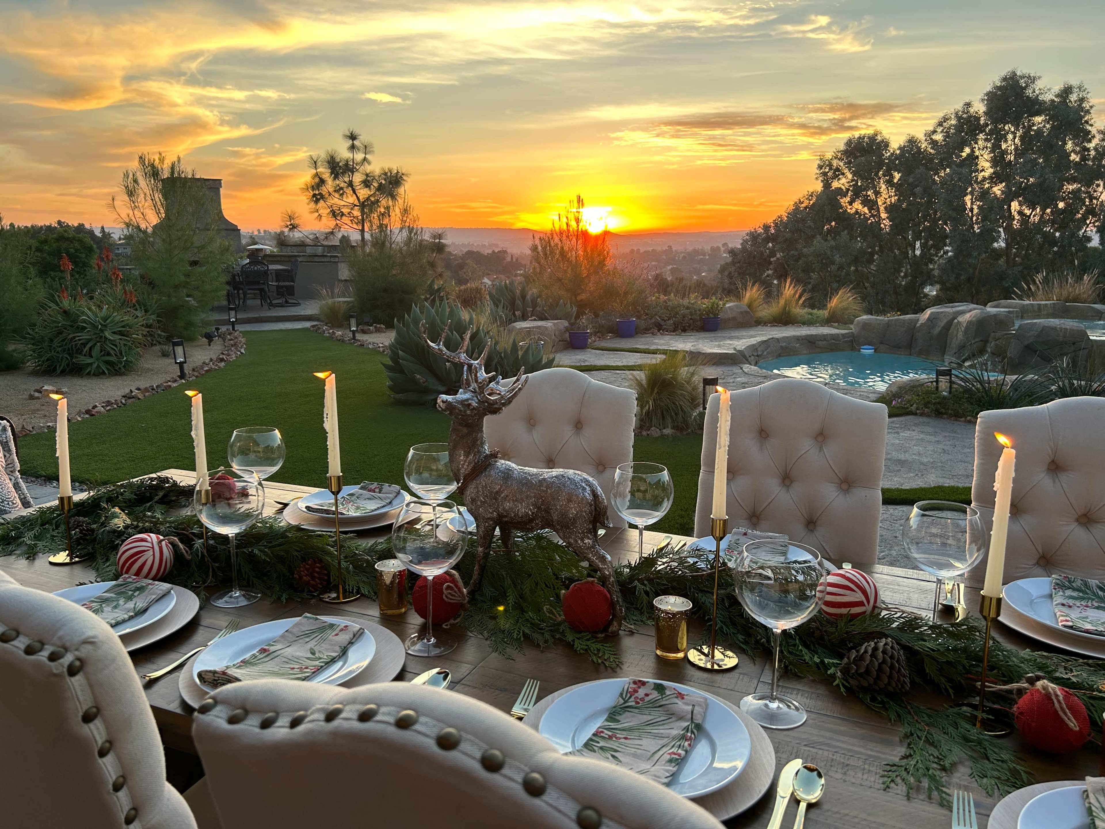 A beautifully set outdoor dining table with candles and decorations overlooks a sunset across a landscaped yard.