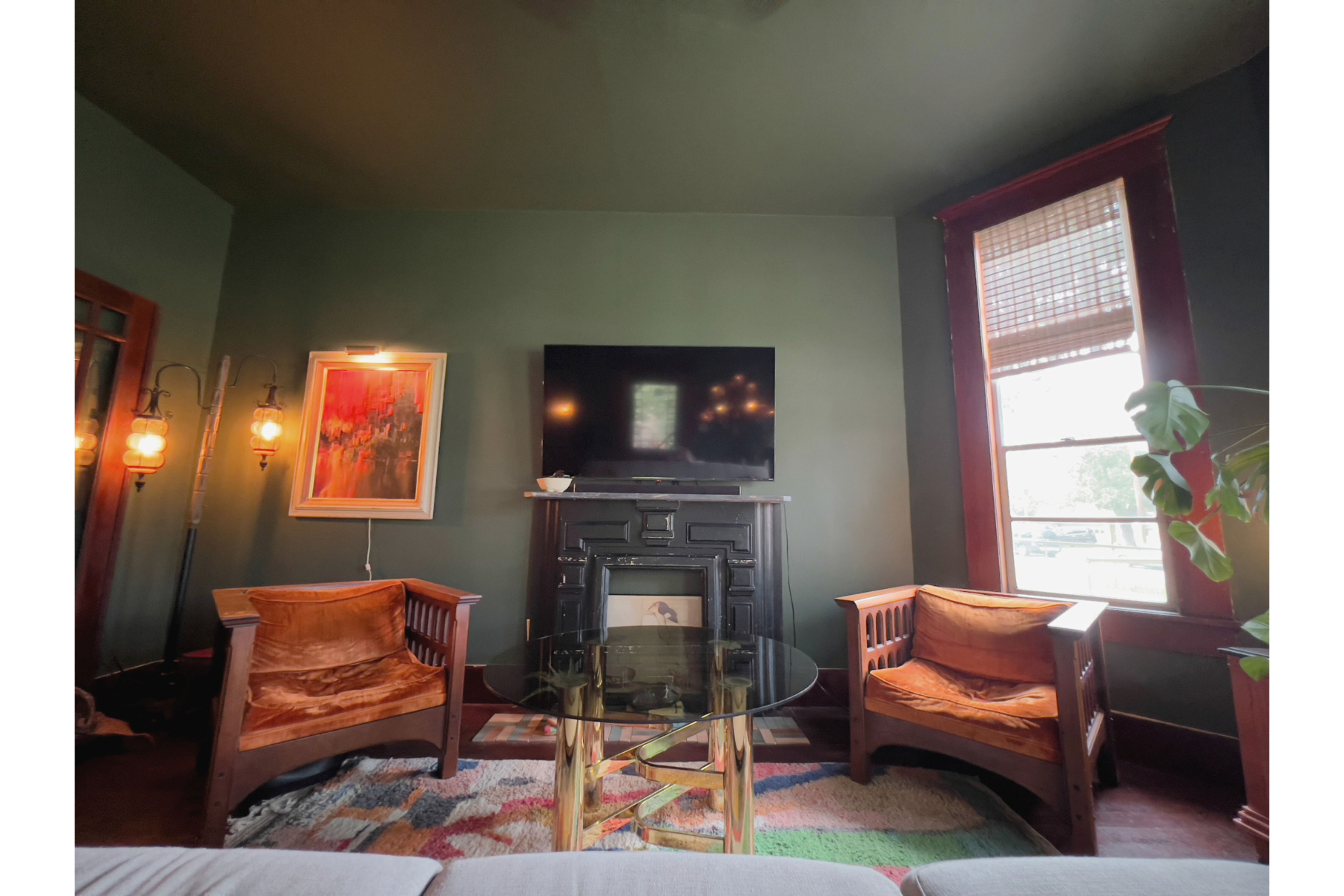 The room features two wooden armchairs facing a glass coffee table, with a television mounted above a decorative fireplace against a green wall.