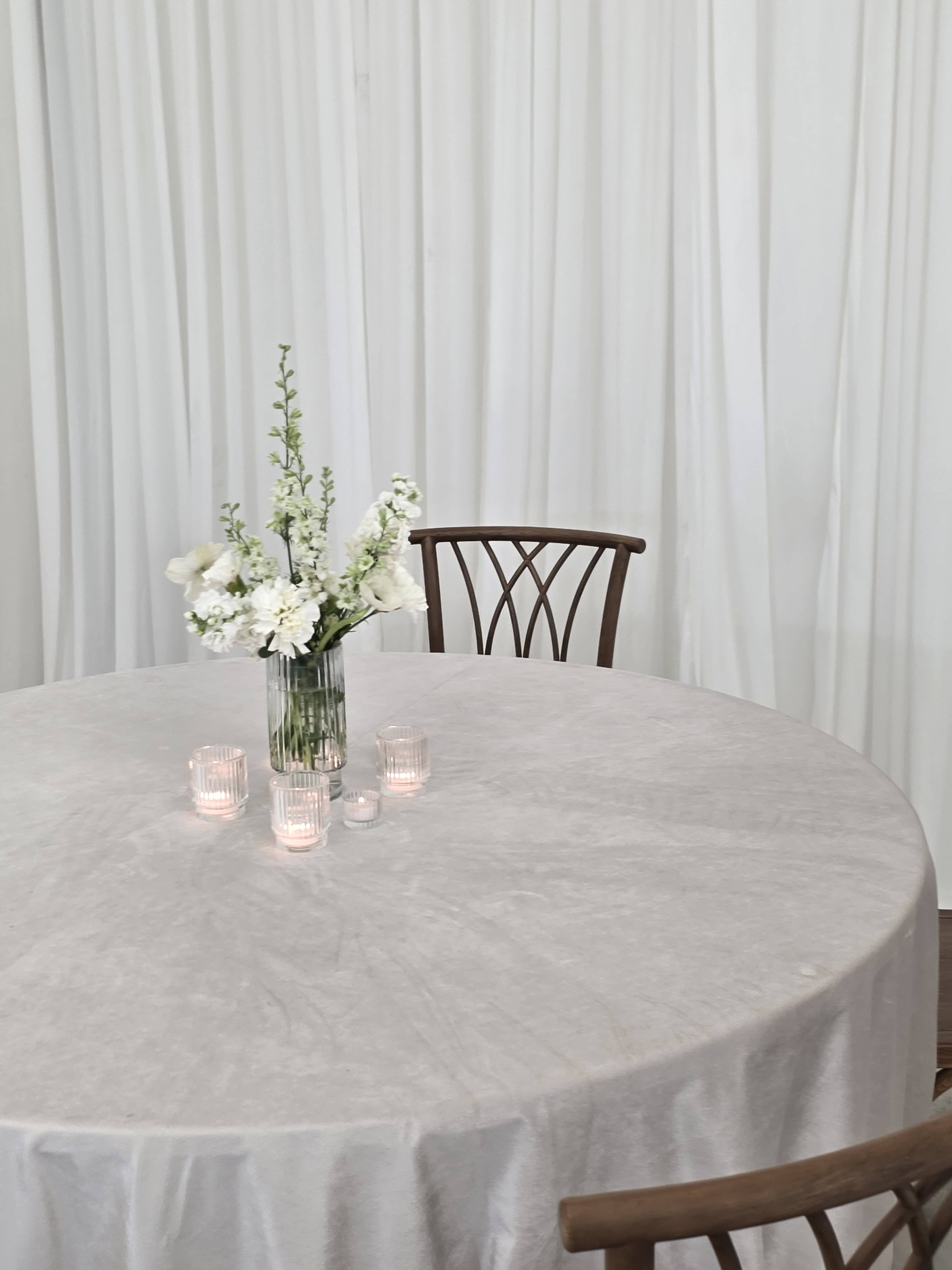 A round table is set with a gray tablecloth, a vase of white flowers, and several tealight candles, against a backdrop of hanging white fabric.
