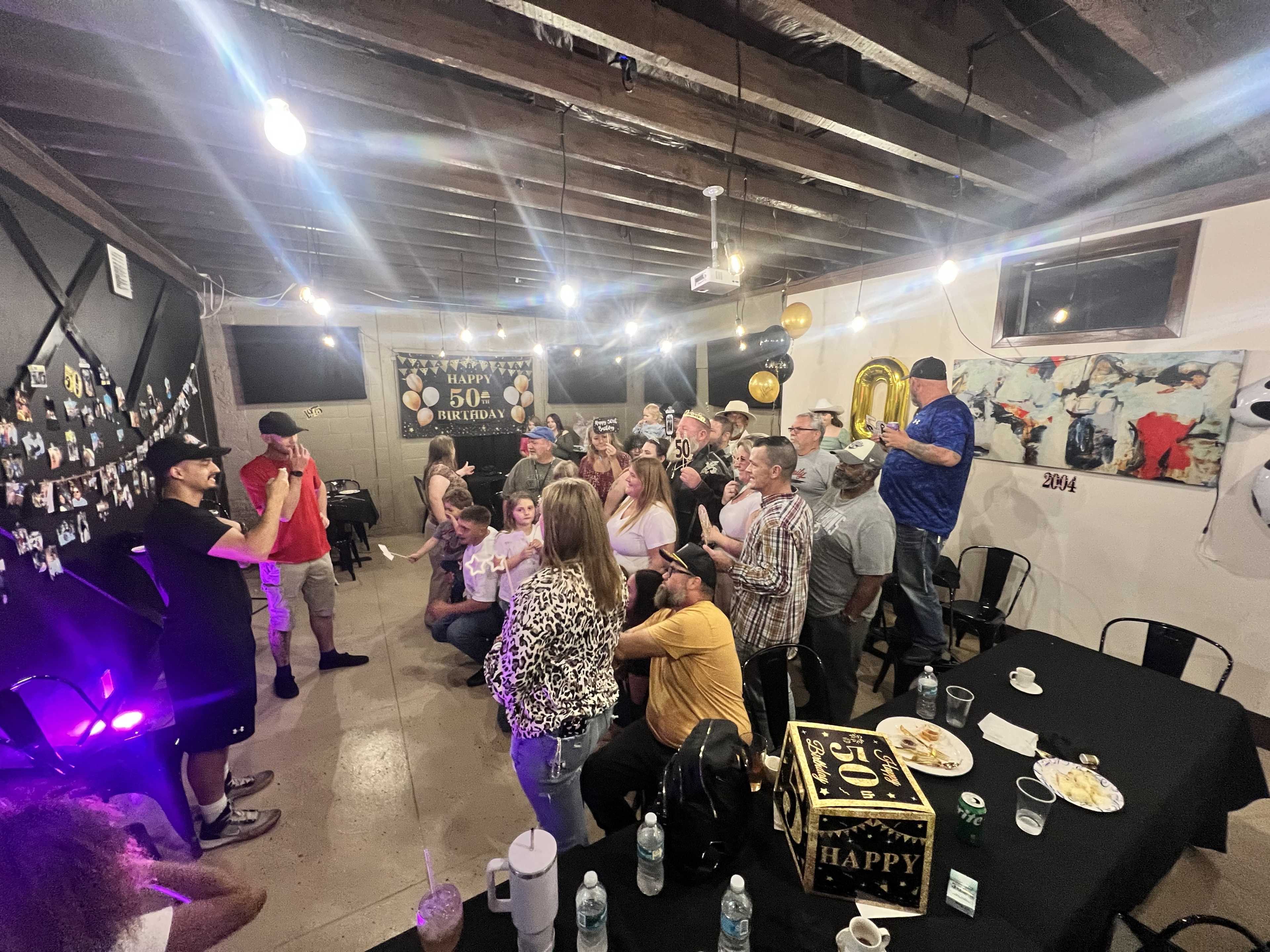 A crowded room celebrates a 50th birthday party with guests gathered around a speaker and decorations visible on the walls.