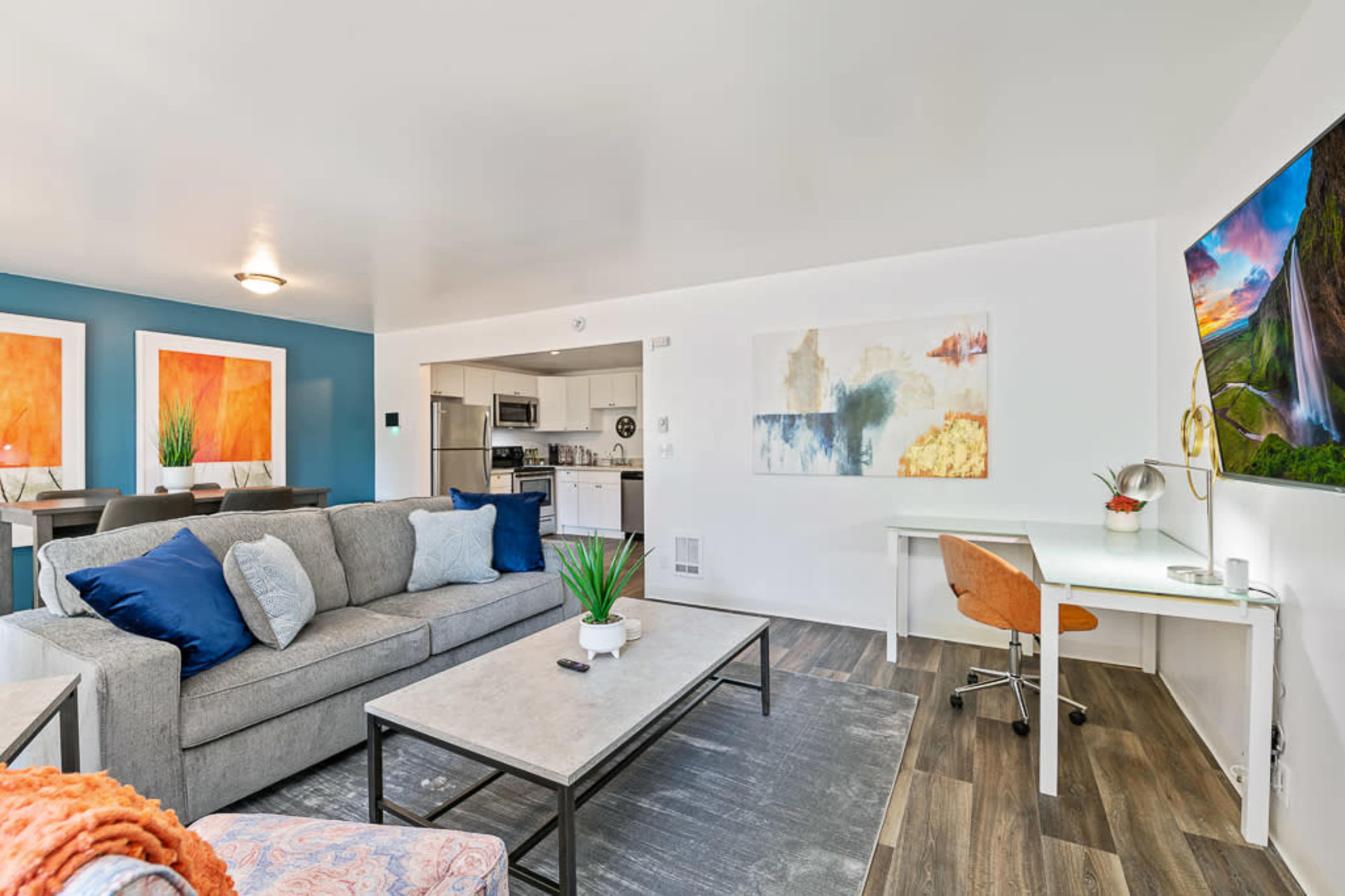 The image shows a modern living room with a gray couch, a coffee table, and a desk, featuring colorful artwork on the walls and a view into a kitchen area.