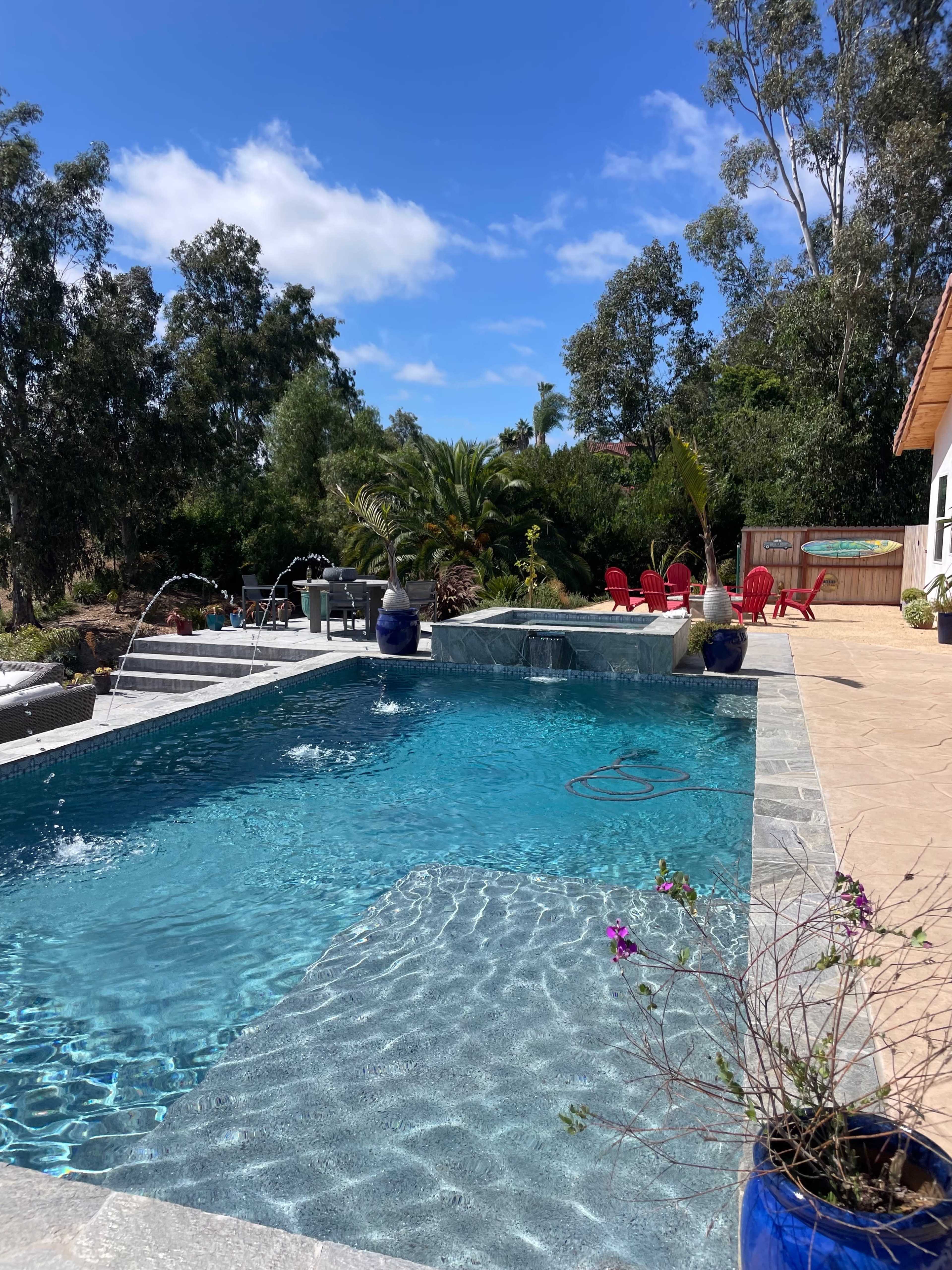 A clear swimming pool surrounded by lush greenery and outdoor furniture, with a fountain feature on one side.