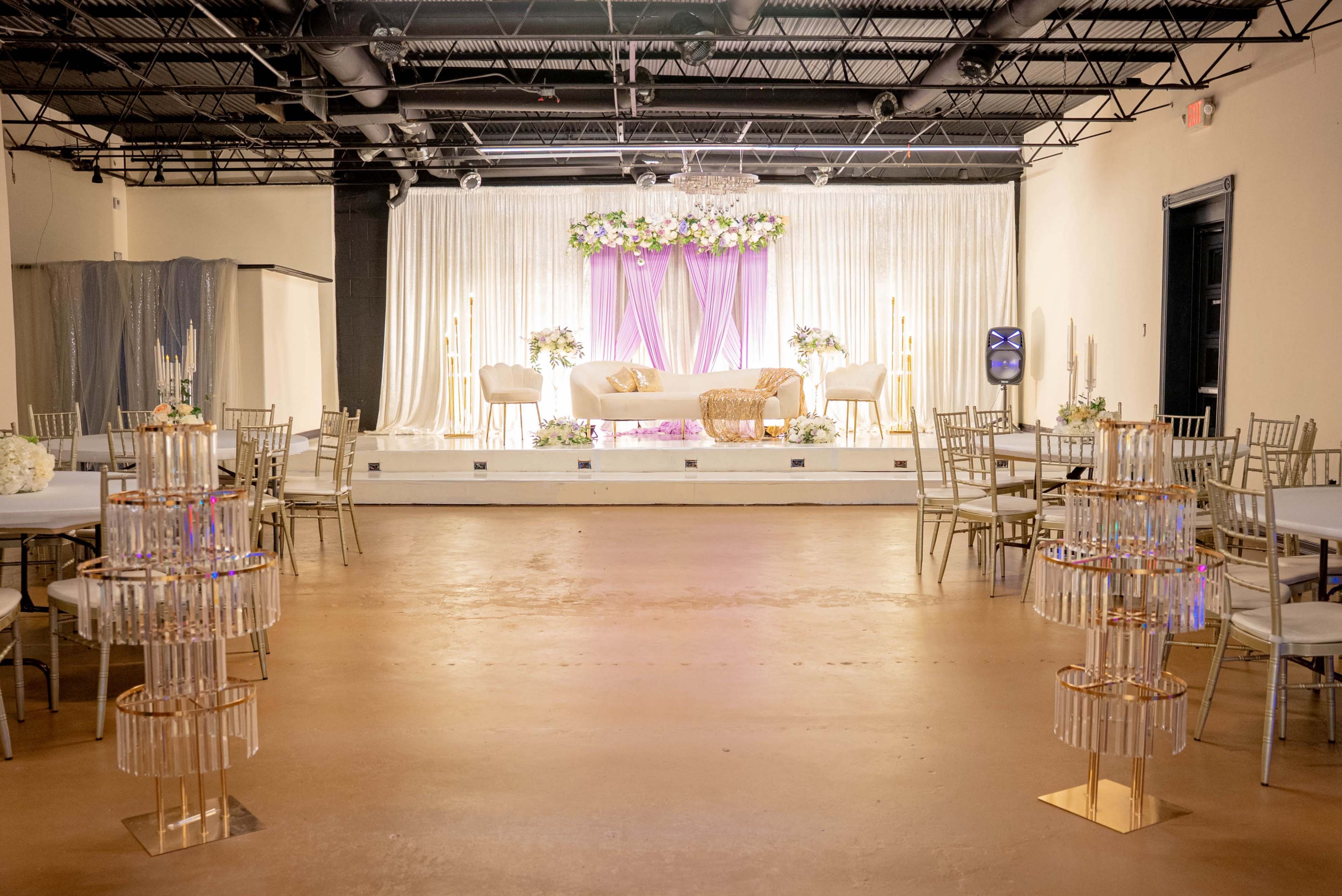 The image shows a spacious event venue featuring a raised stage decorated with floral arrangements and elegant seating, surrounded by tables and transparent chairs.