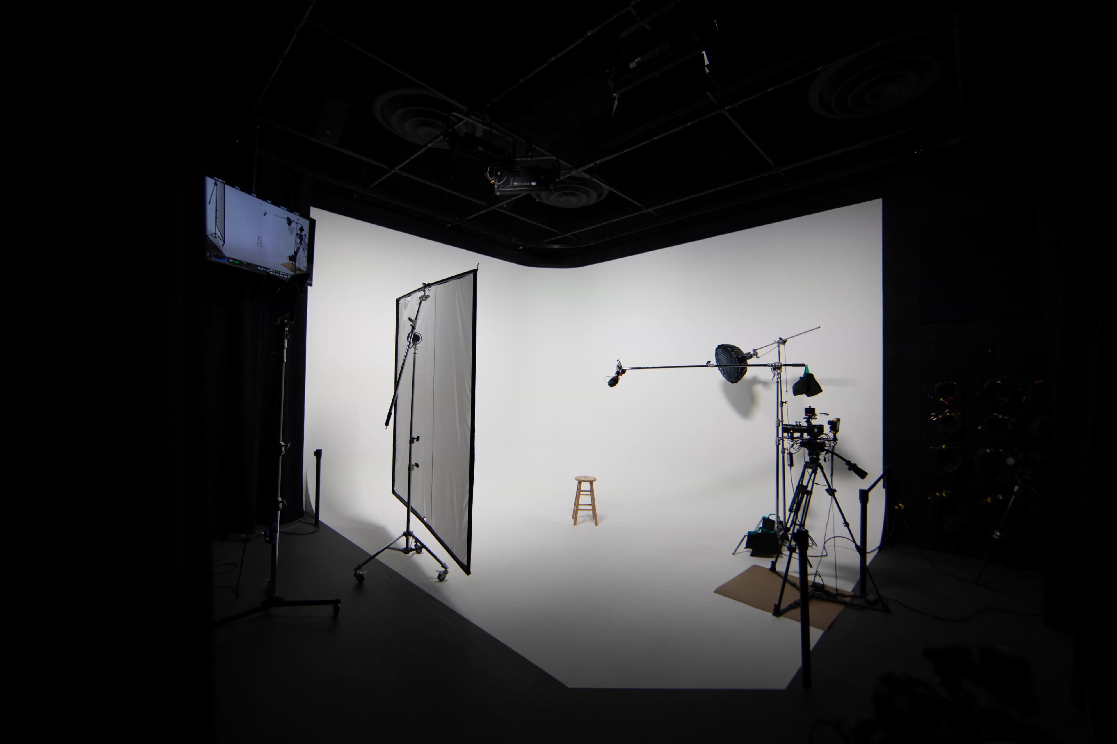 A photography studio featuring a white backdrop, a stool in the center, and various lighting and camera equipment arranged around the space.