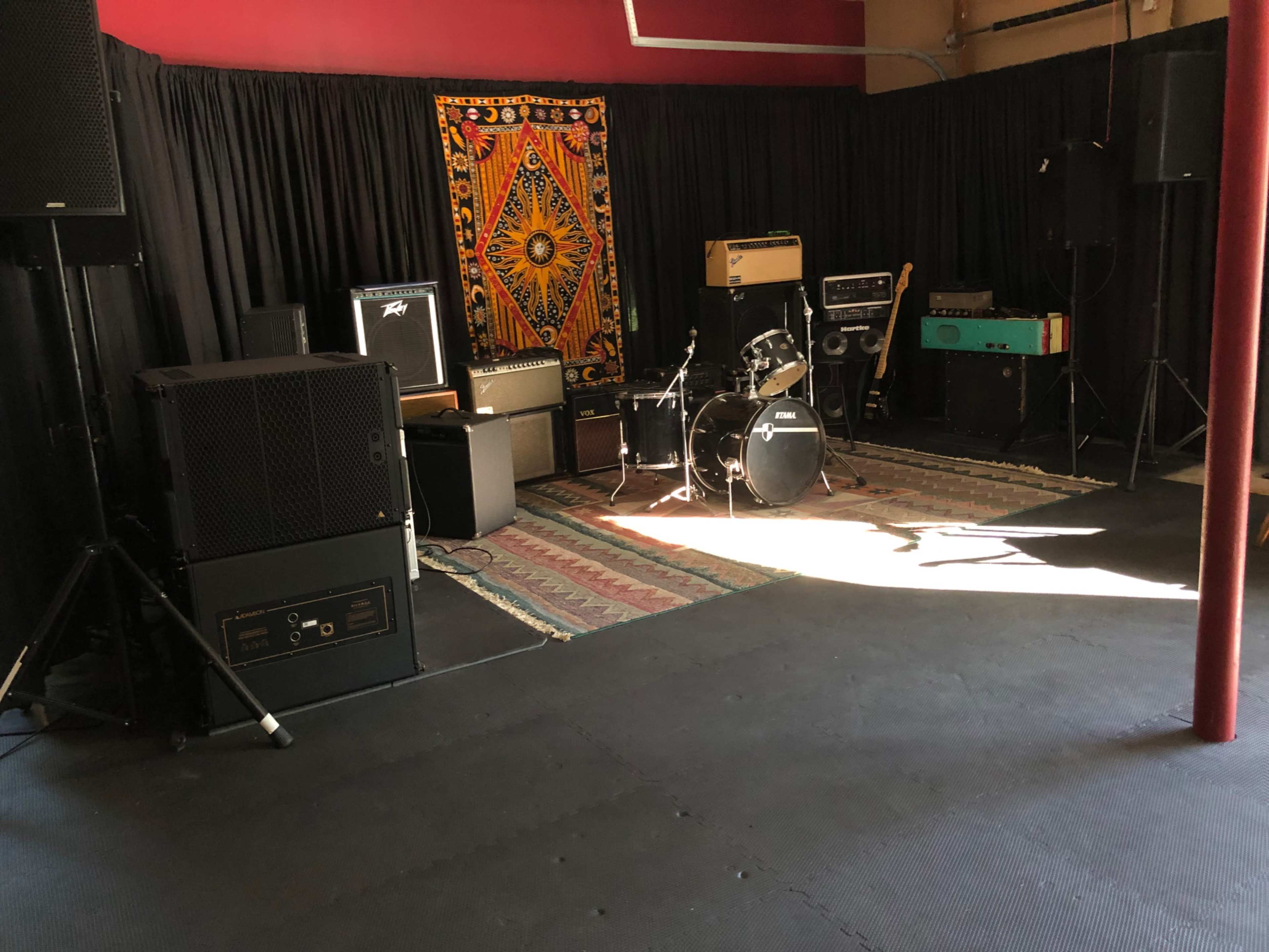 The image shows a music rehearsal space with various amplifiers, a drum set, and a colorful tapestry hanging on the wall.