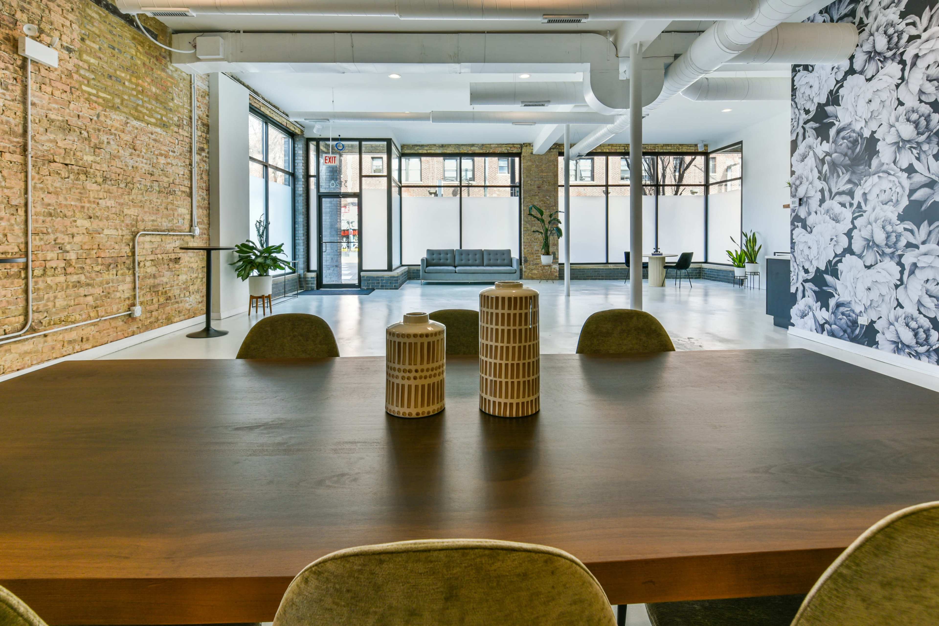 A spacious modern interior with a wooden dining table in the foreground and large windows that provide a view of an adjacent seating area and floral wallpaper on one wall.