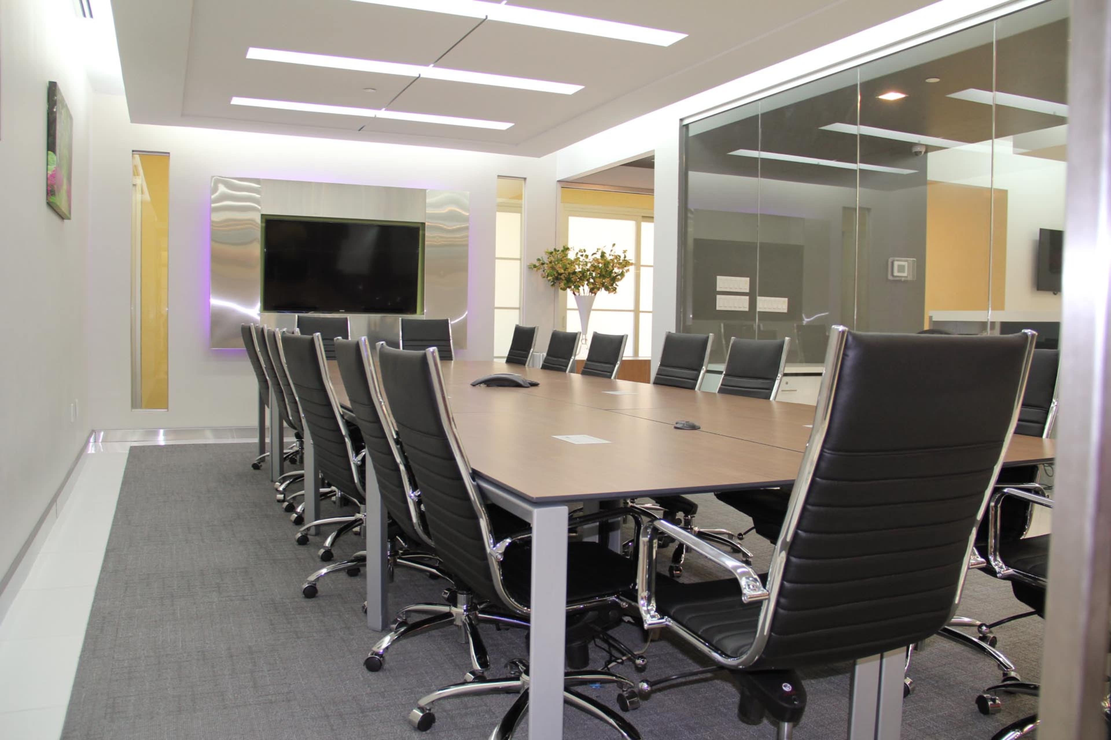 A spacious conference room features a long wooden table surrounded by black ergonomic chairs, with a large television screen mounted on the wall.