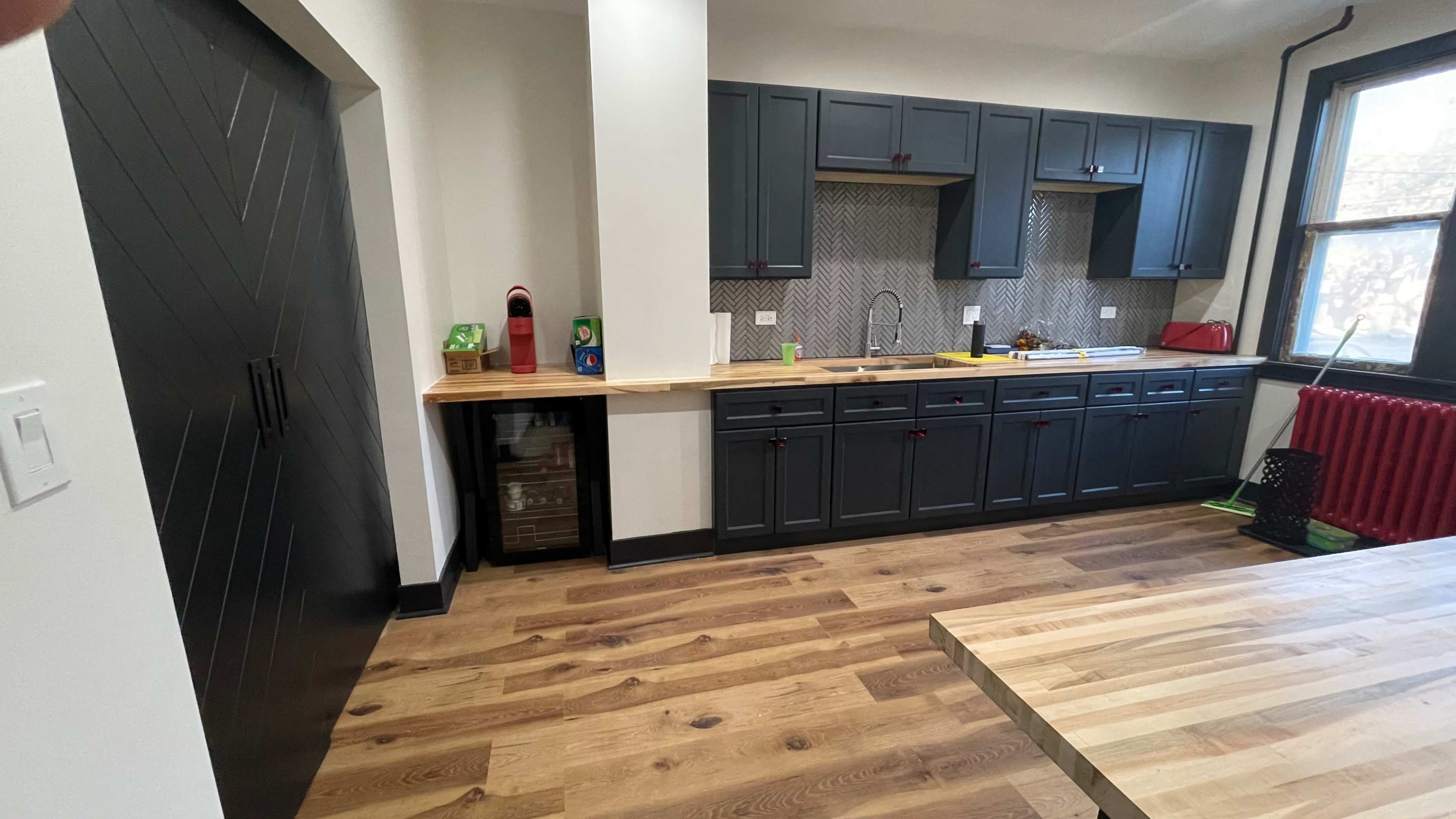 The image shows a modern kitchen with dark cabinetry, a wooden countertop, and a small beverage refrigerator beside a pantry door.