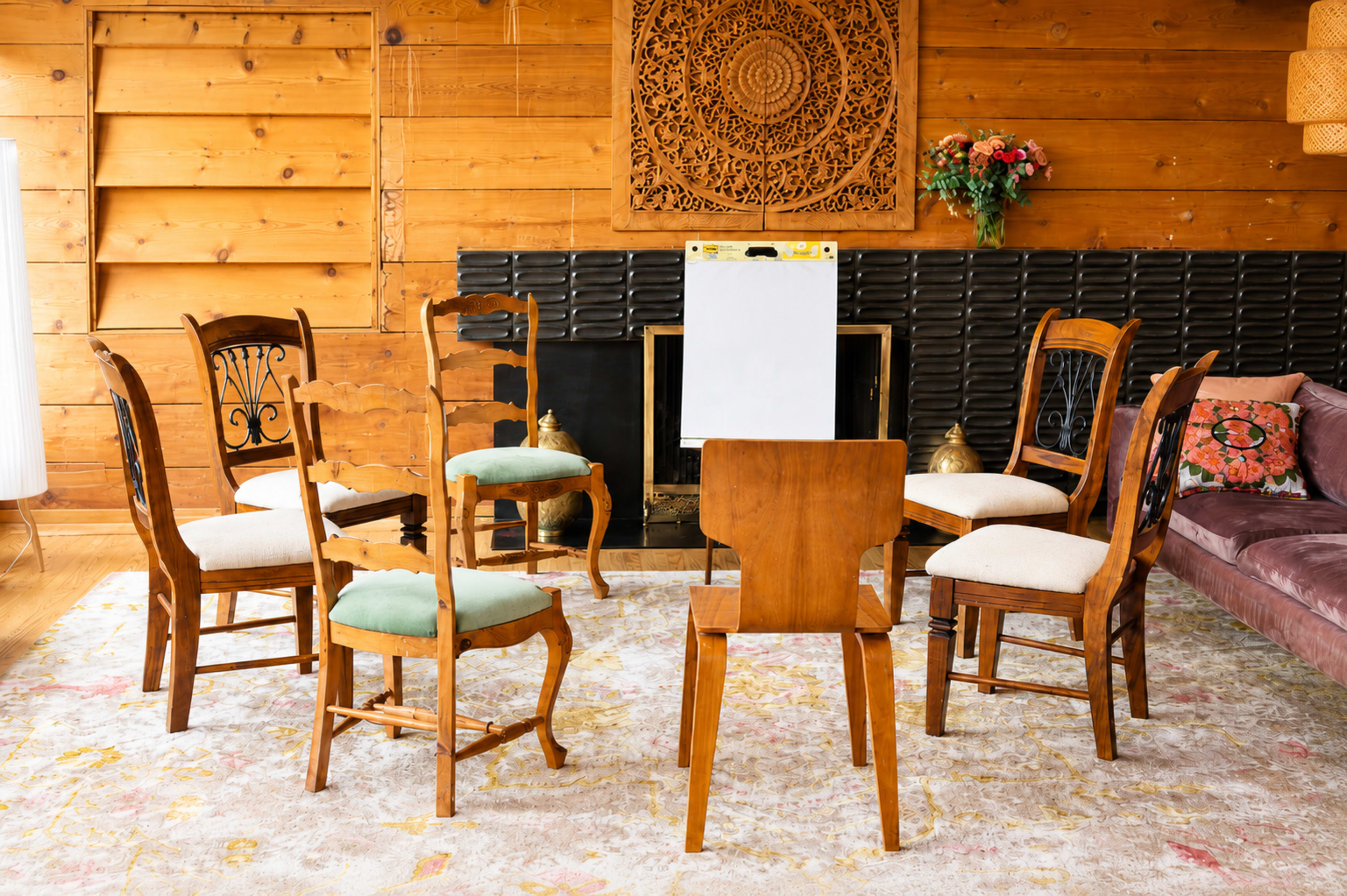 The image shows a circle of wooden chairs arranged around a blank easel in a cozy, rustic room with wooden walls and patterned rugs.