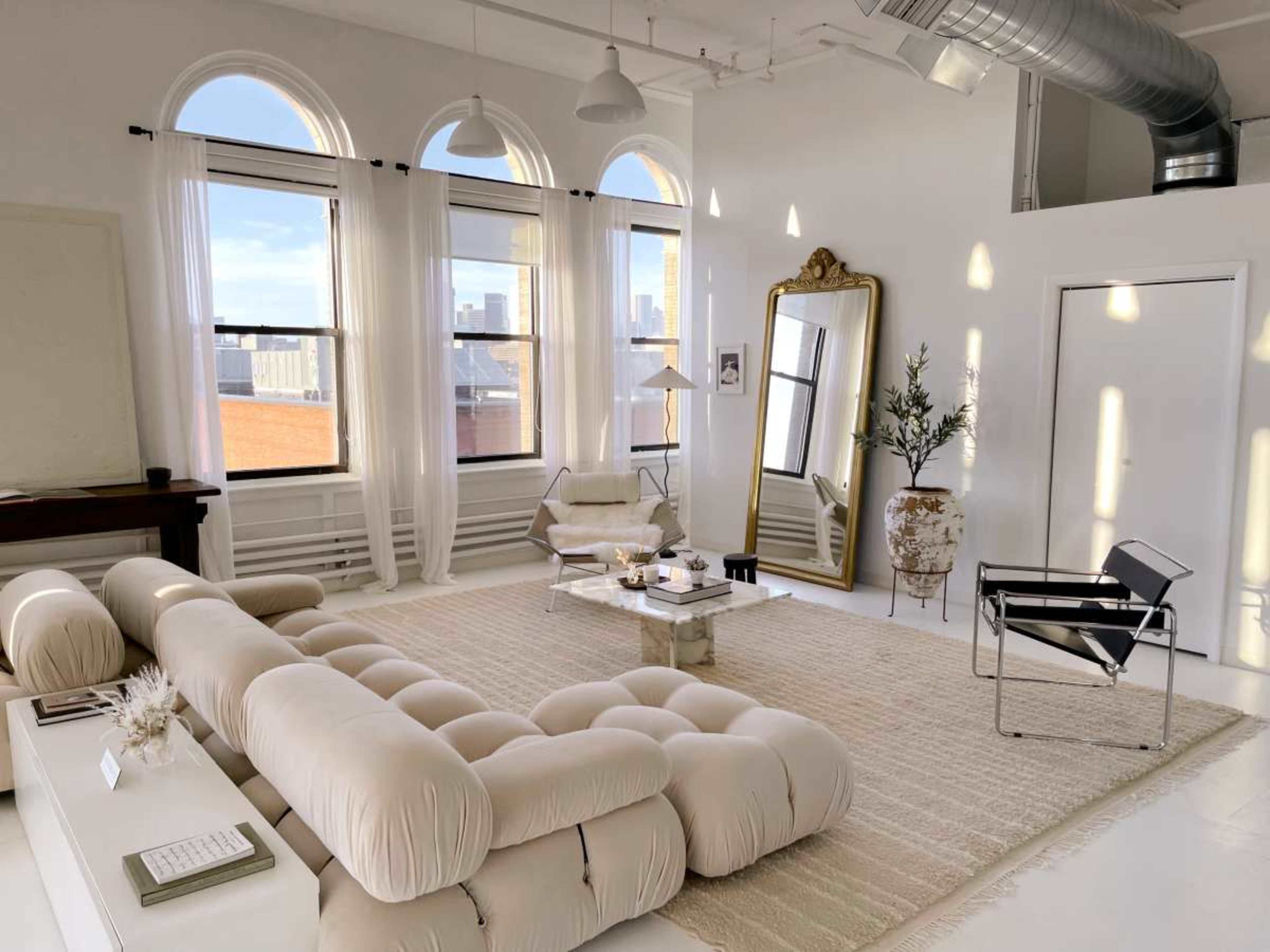 The image shows a brightly lit, spacious interior with large windows, featuring a light-colored, tufted sofa, a decorative mirror, a small coffee table, and minimalist decor arrangements.