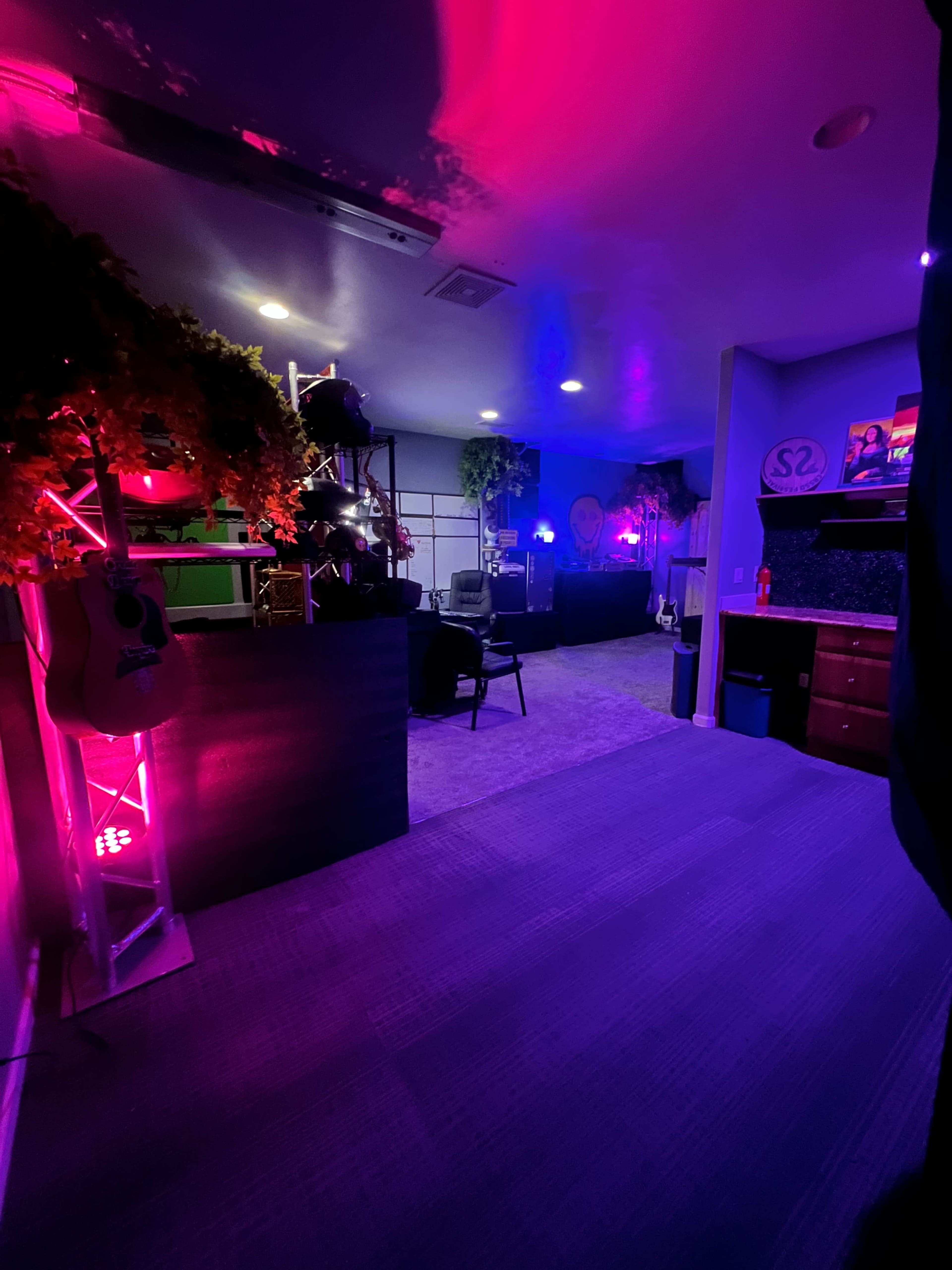 The image shows a dimly lit room featuring colorful lighting, musical instruments, and a seating area.