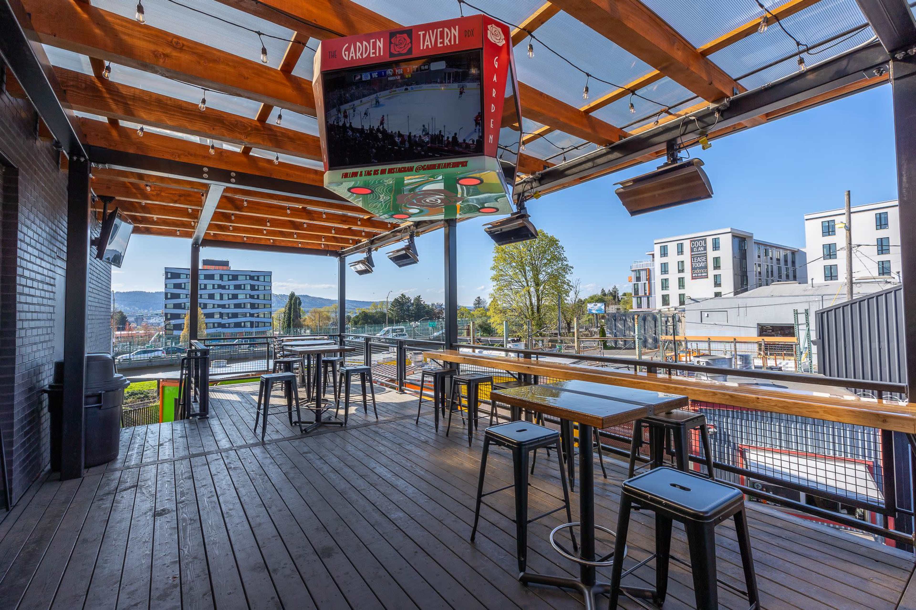 An outdoor patio area of a bar or restaurant with wooden tables and metal stools, featuring a large screen mounted above for viewing events.