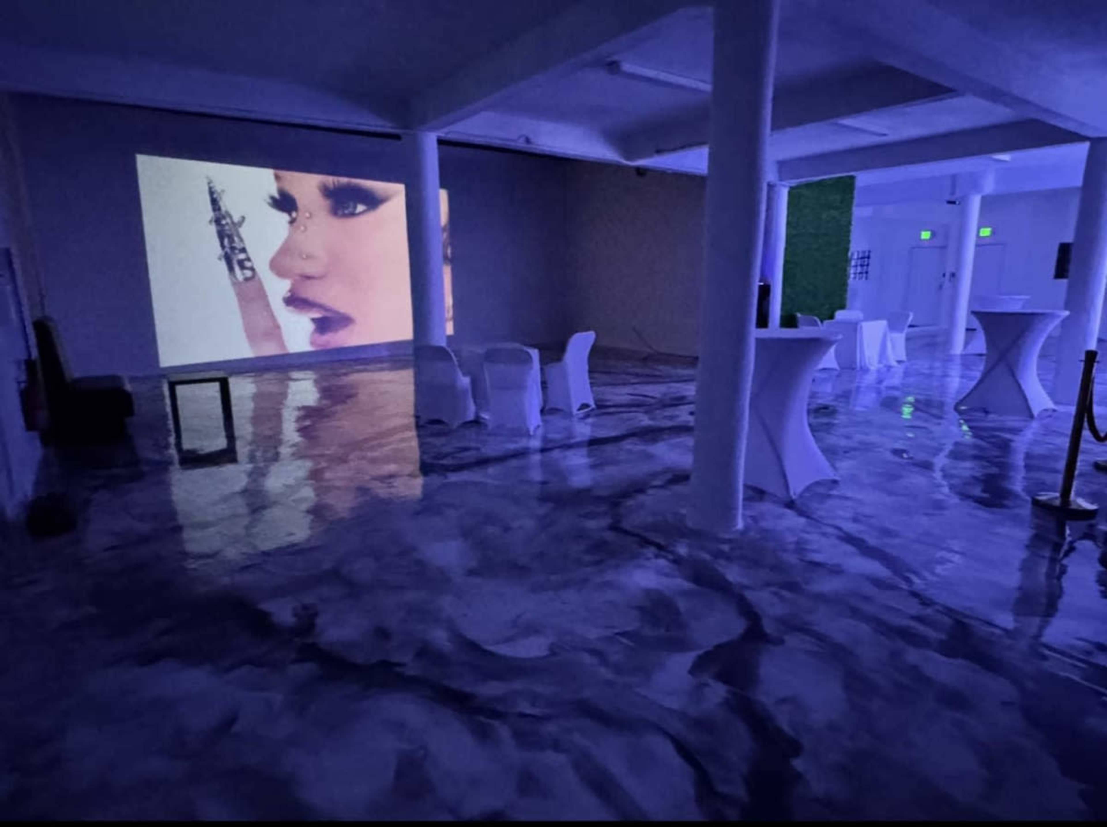 The image shows a dimly lit event space with a large projection on one wall and white tables arranged around a shiny, reflective floor.