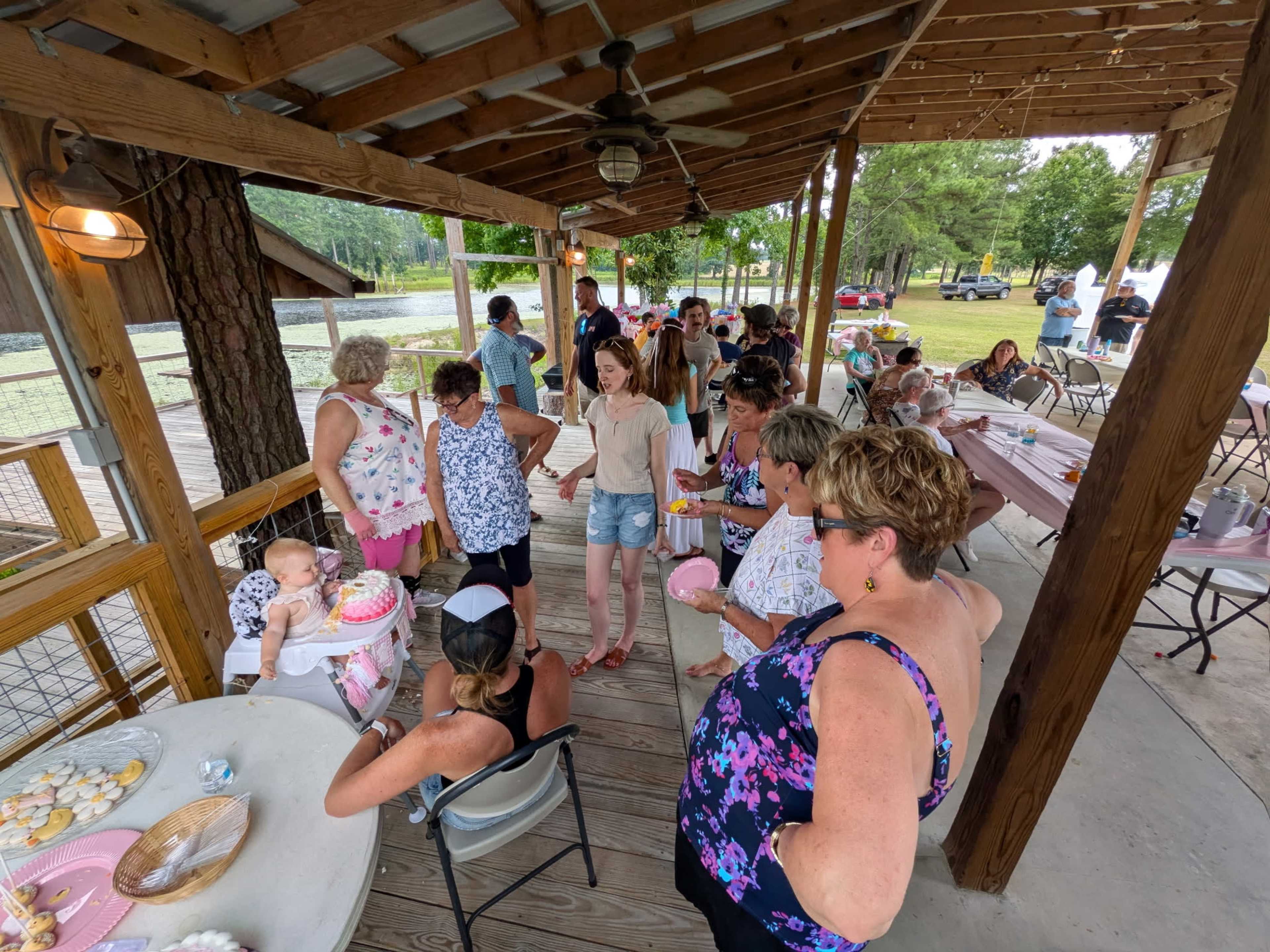 A large group of people gathers under a covered pavilion, socializing and celebrating at a party with tables set up for food and drinks.