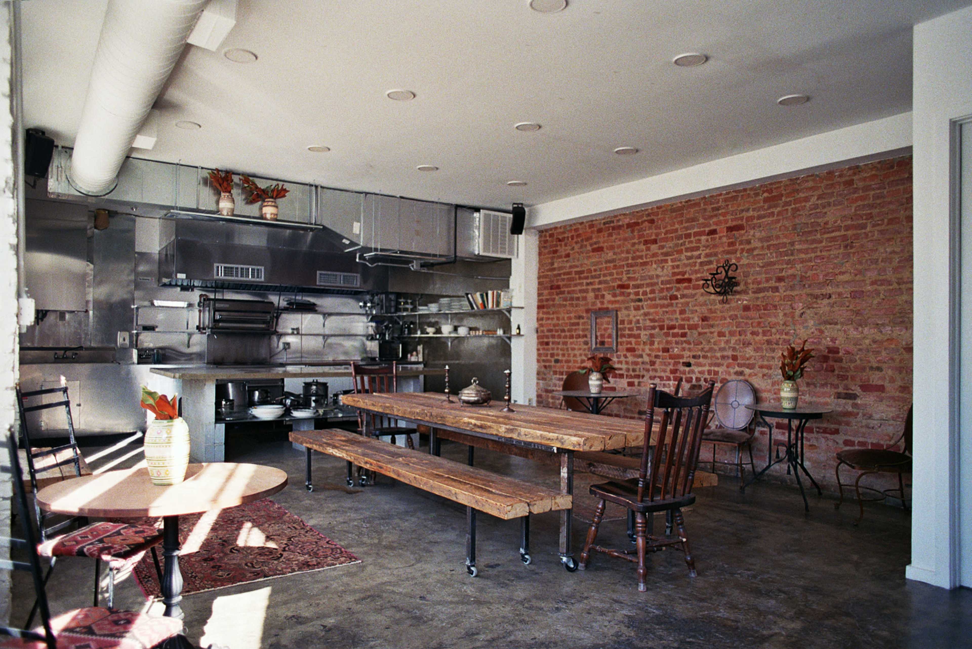 The image shows a minimalistic restaurant interior featuring a brick wall, wooden tables, and a kitchen area with stainless steel appliances.