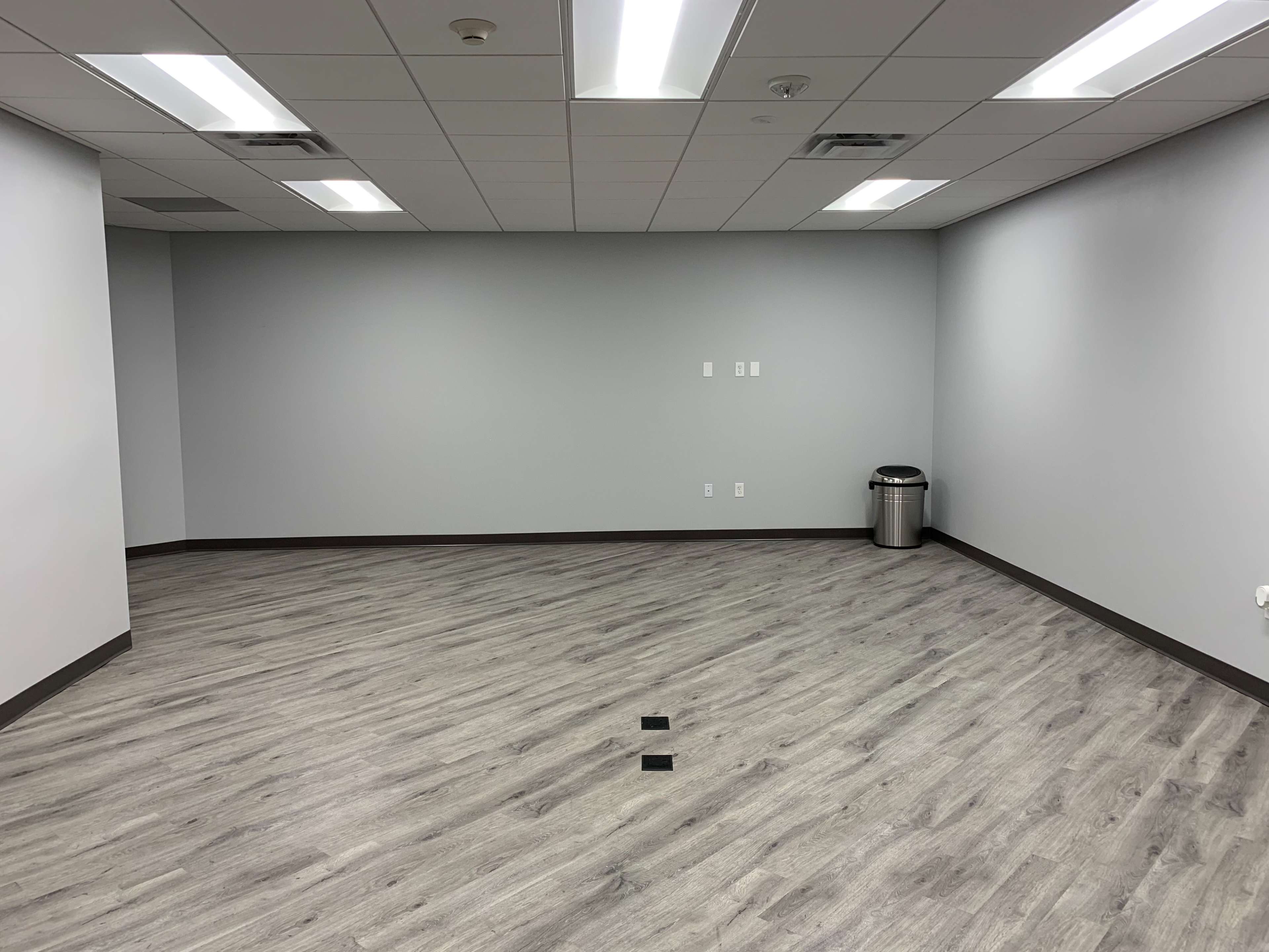 The image shows a blank office room with gray walls, a trash can in the corner, and light-colored flooring.