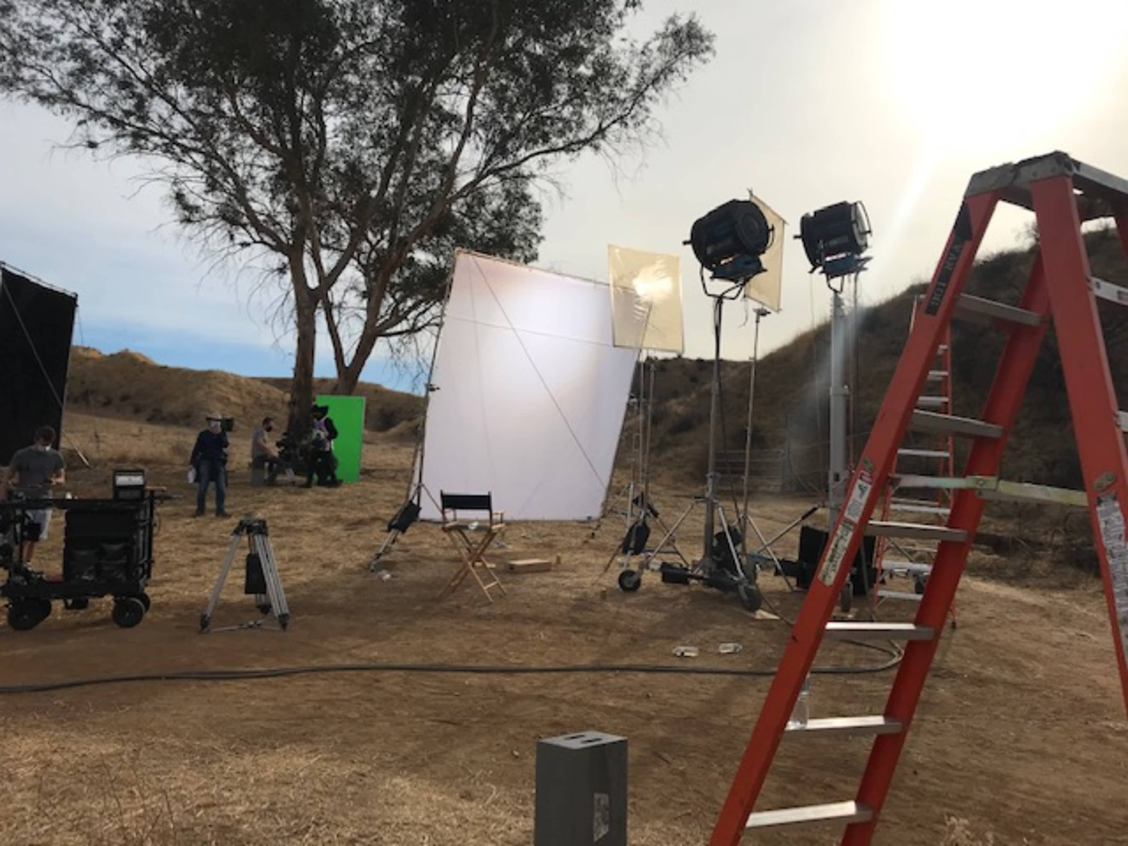 A film set is arranged outdoors with lighting equipment, a green screen, and a ladder positioned next to a tree on a dry hillside.