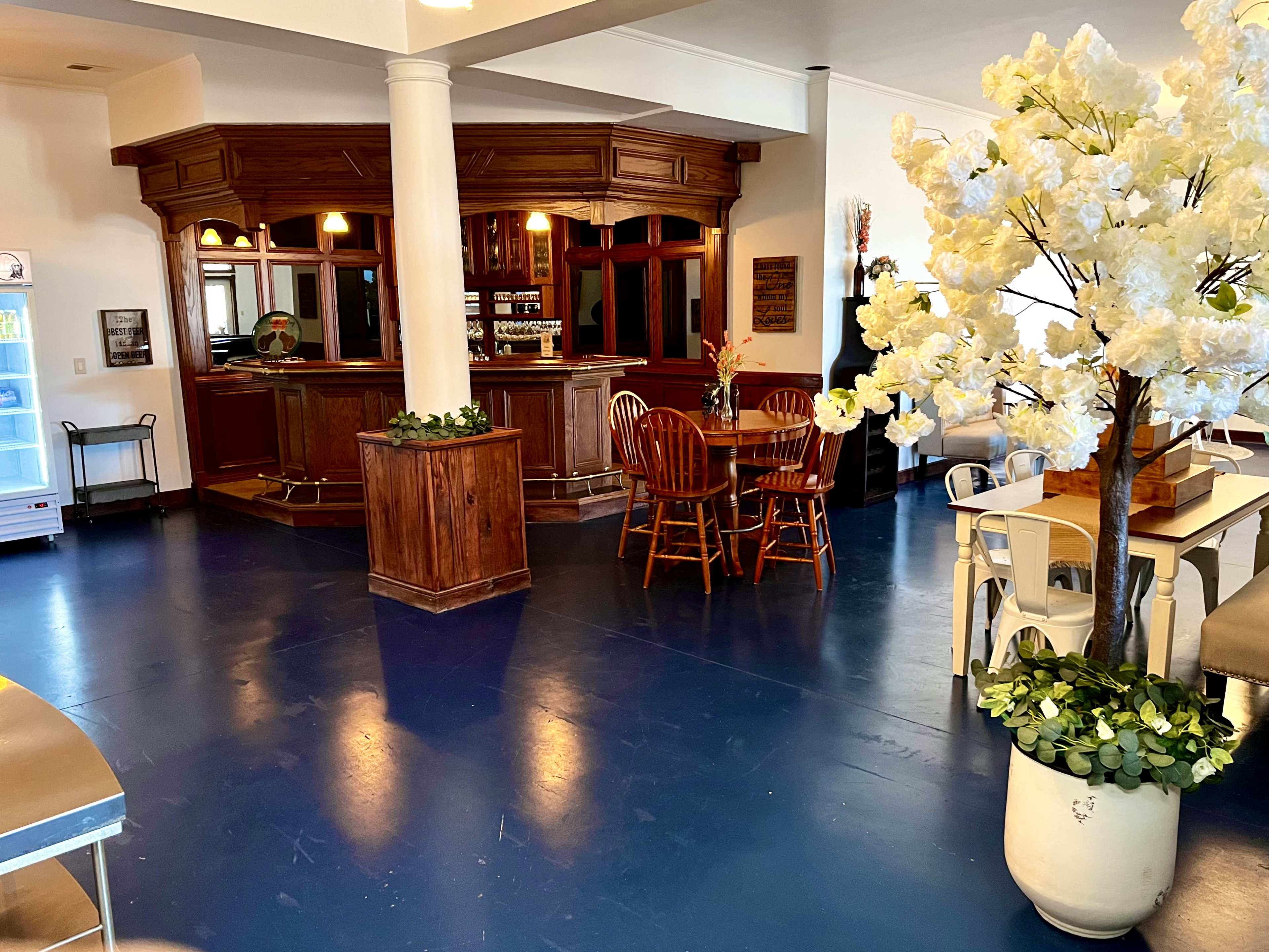 The image shows a spacious interior with a bar area, wooden seating, and a decorative flower arrangement.