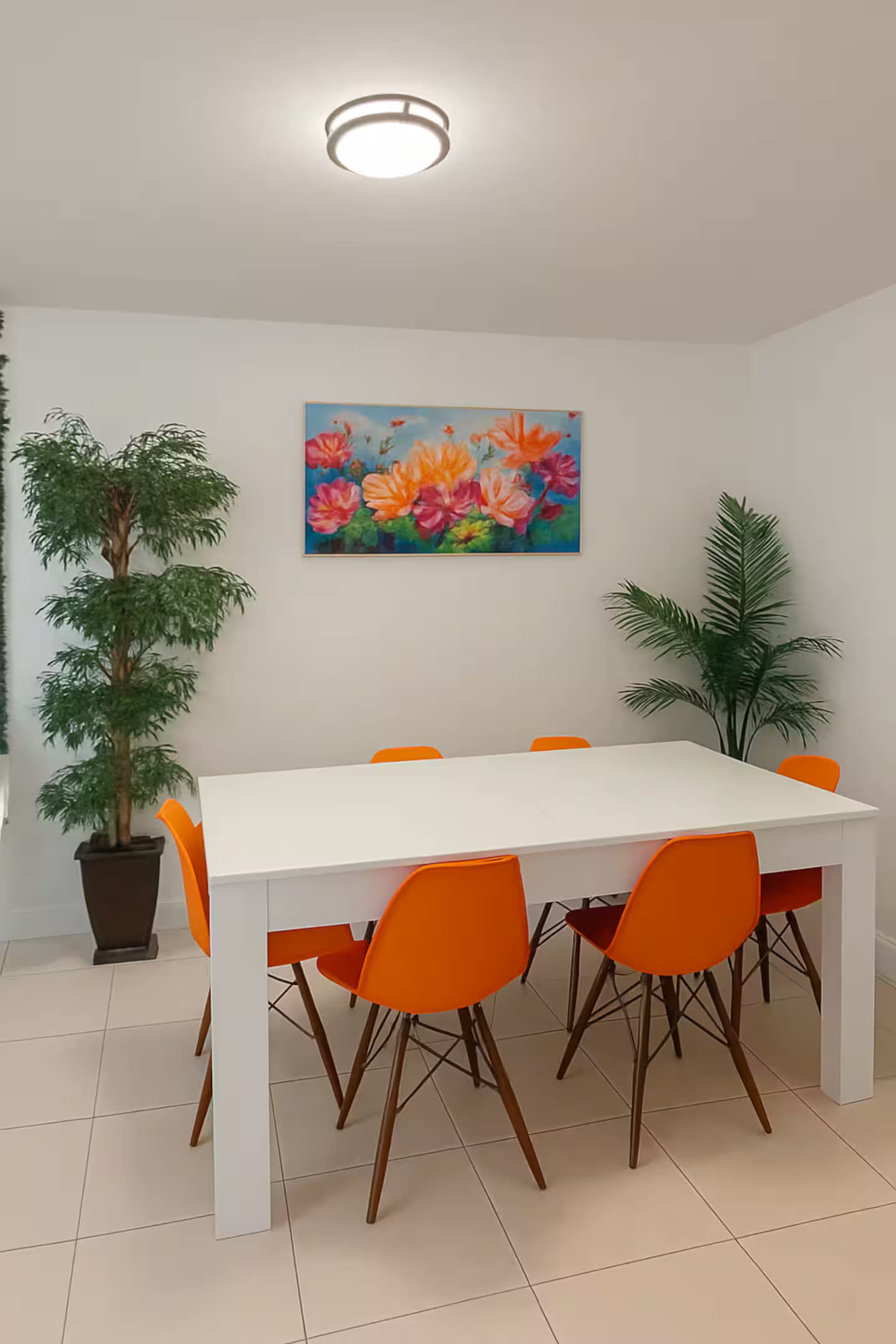 A bright dining area features a white table surrounded by orange chairs, with a floral painting on the wall and two potted plants nearby.