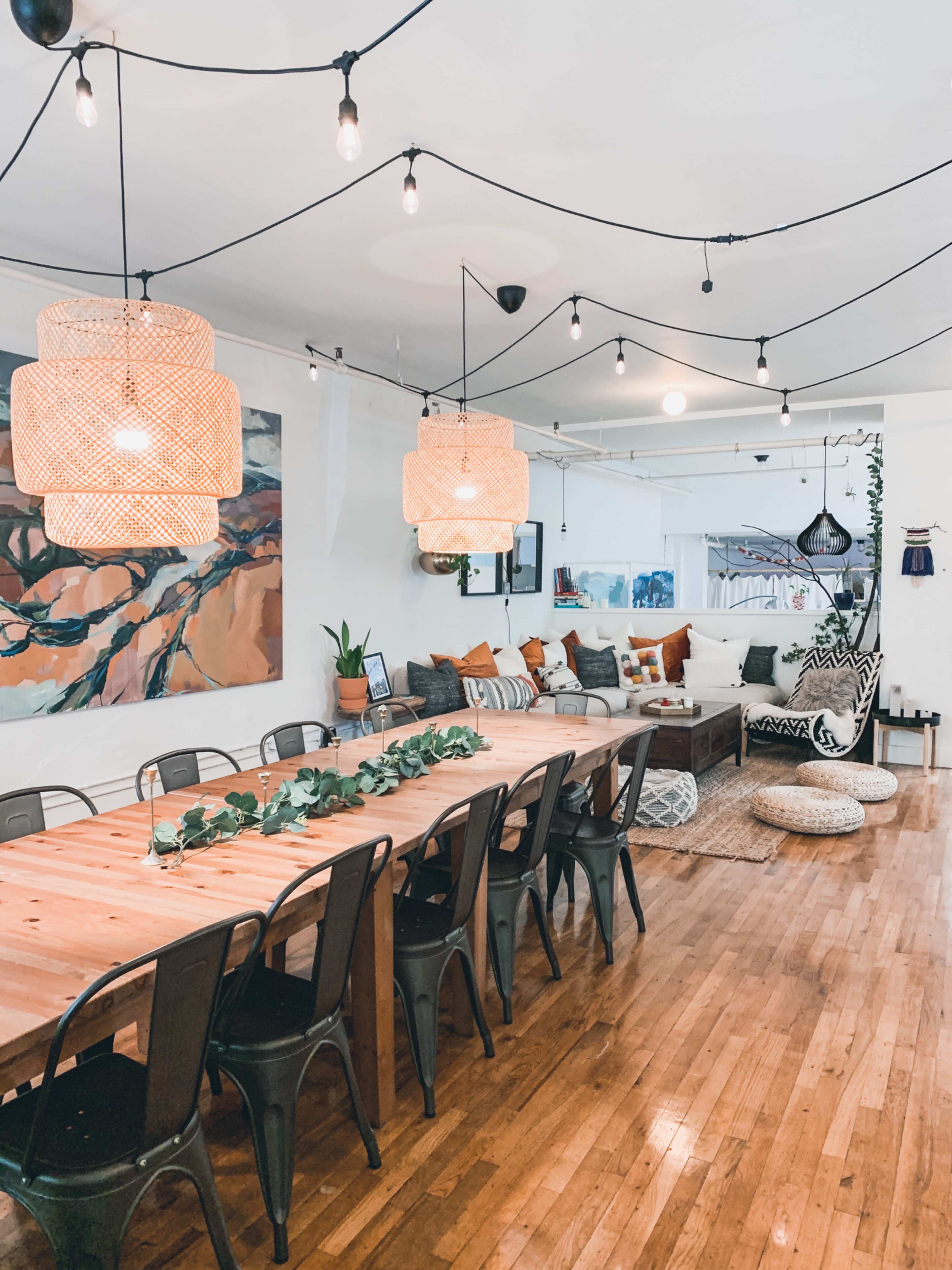 A spacious dining area featuring a long wooden table with metal chairs, pendant lights hanging overhead, and a cozy seating area with cushions and artwork on the walls.