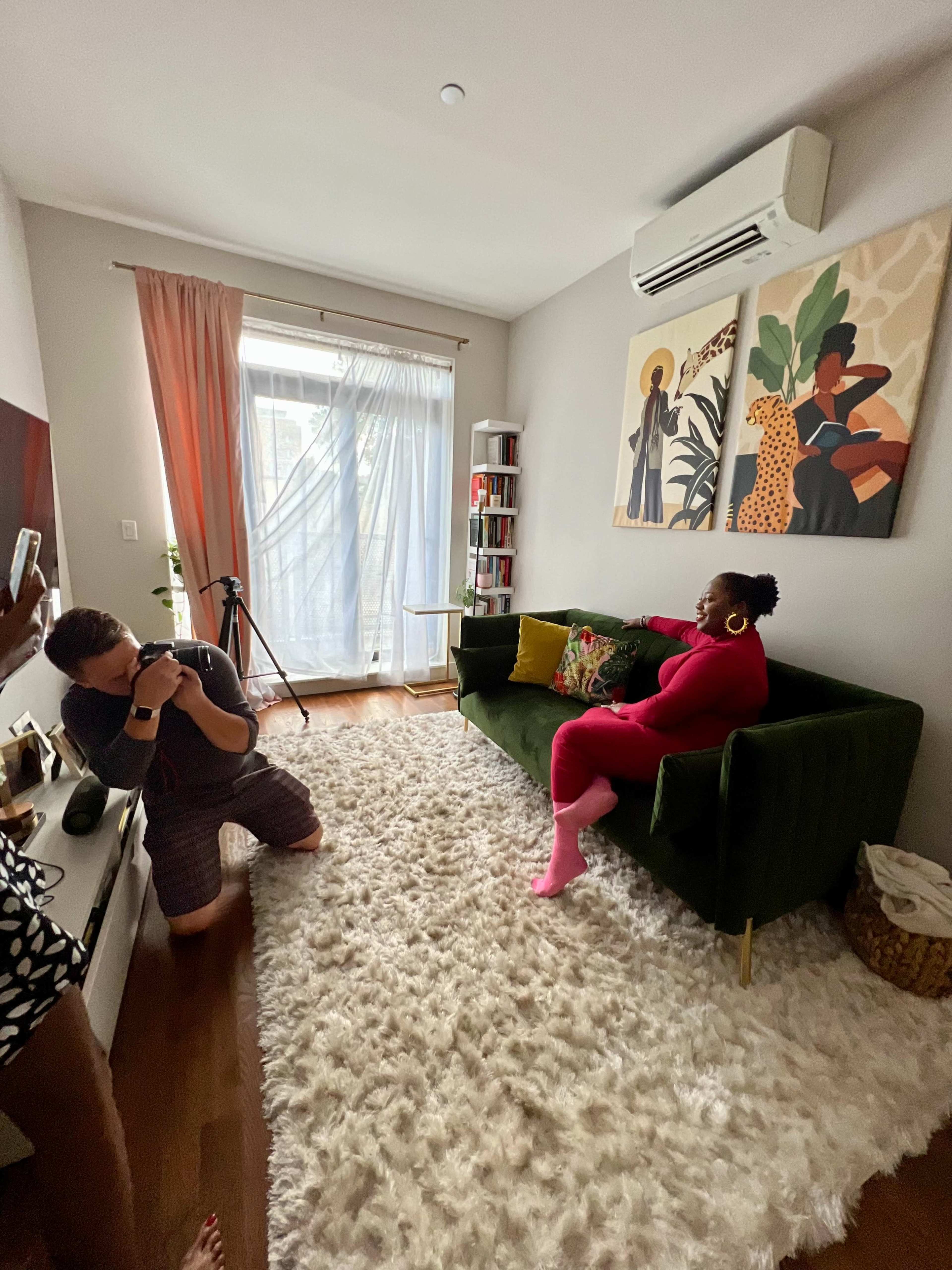 A woman sits on a green couch in a living room while a person kneels nearby, taking photos.