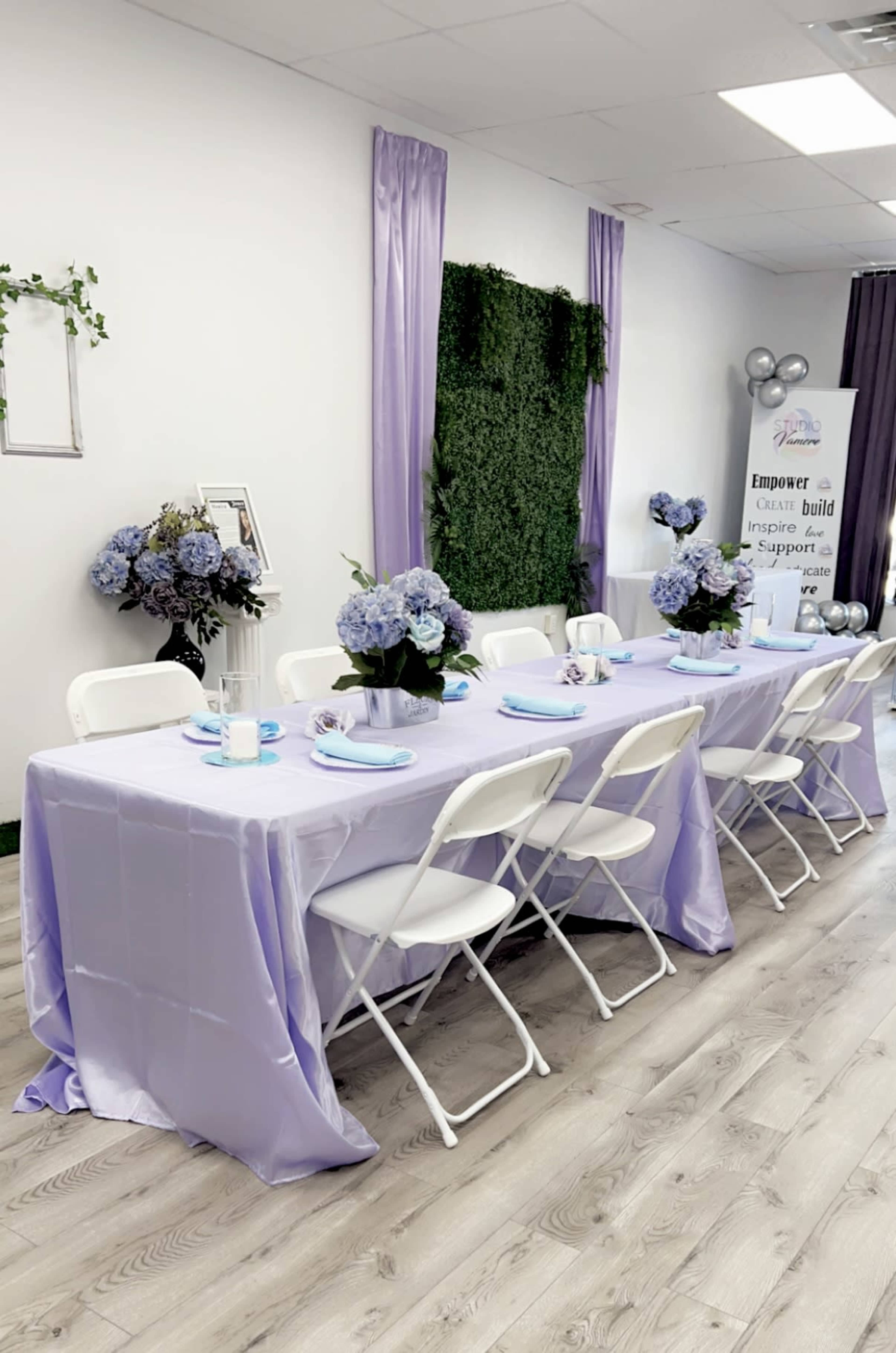 A table set for an event, covered with a light purple tablecloth and surrounded by white chairs, features decorative floral arrangements and a banner promoting empowerment and support.