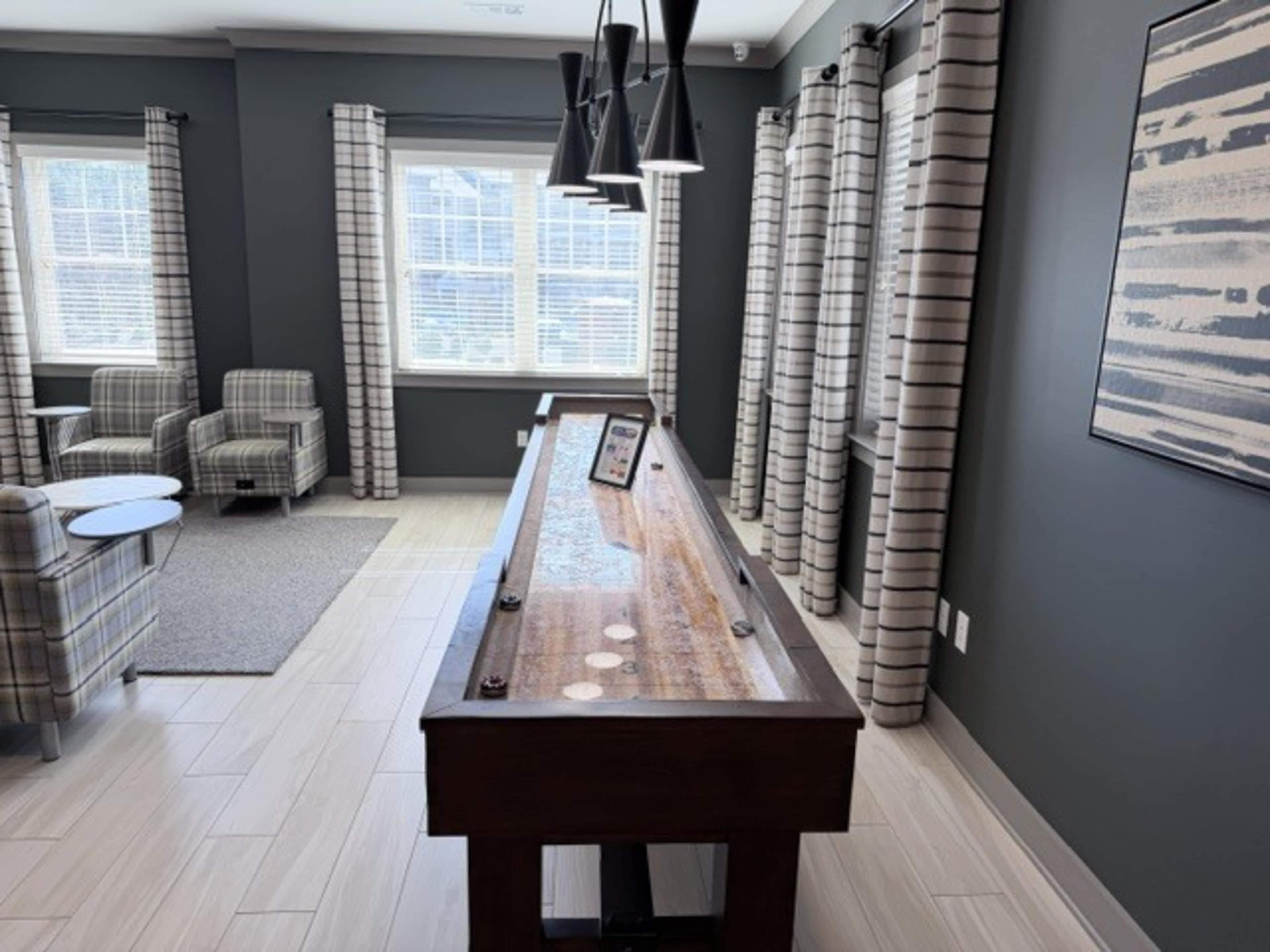 The image shows a game room featuring a shuffleboard table, with two striped curtains and comfortable chairs in the background.