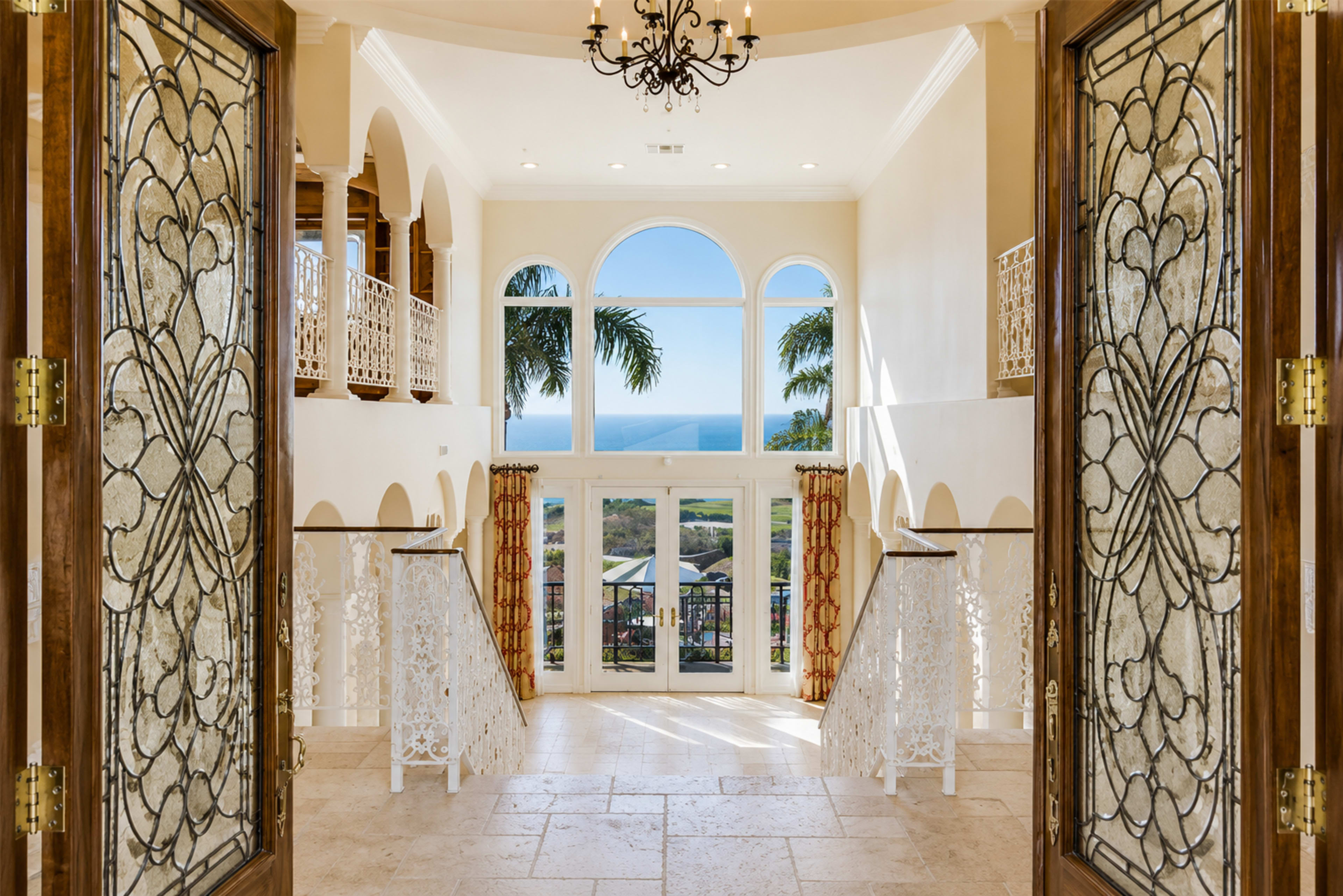 The entrance opens to a spacious hall with a chandelier, flanked by arched windows offering a view of the ocean.