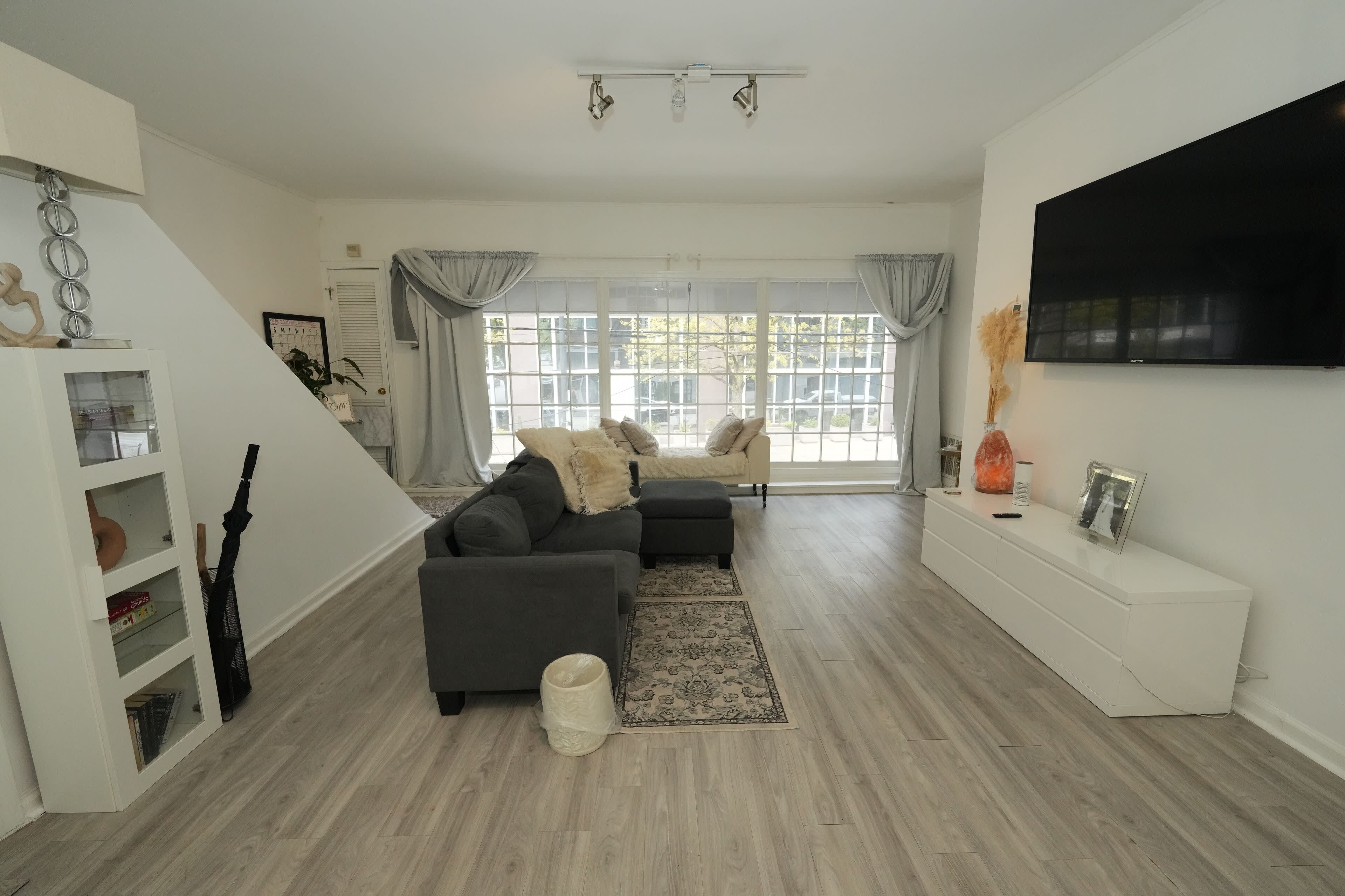 The living room features a gray sectional sofa, a white media console, and large windows with sheer curtains, creating a bright and open atmosphere.