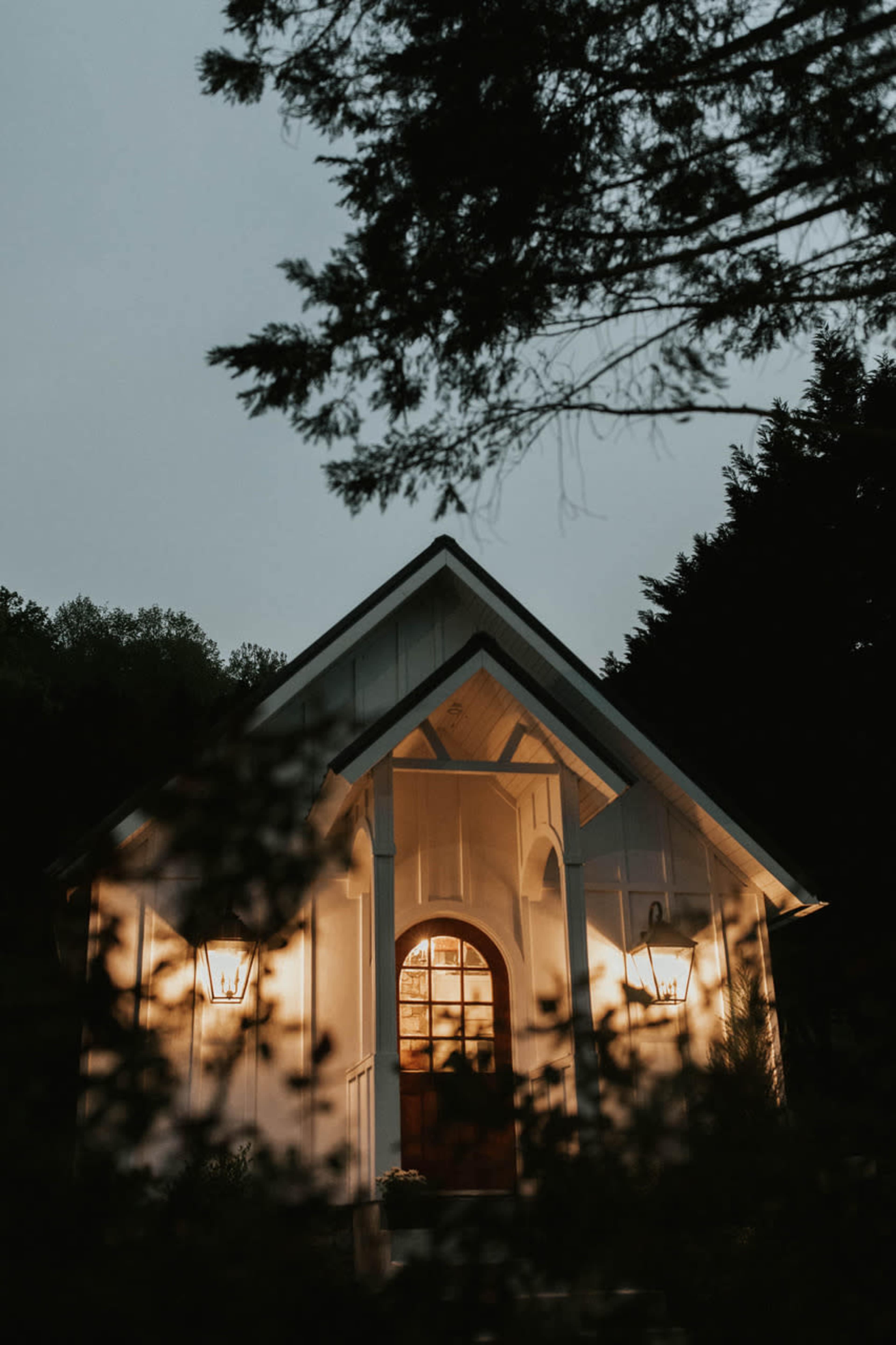 A small white house with illuminated lamps stands amidst trees during twilight.