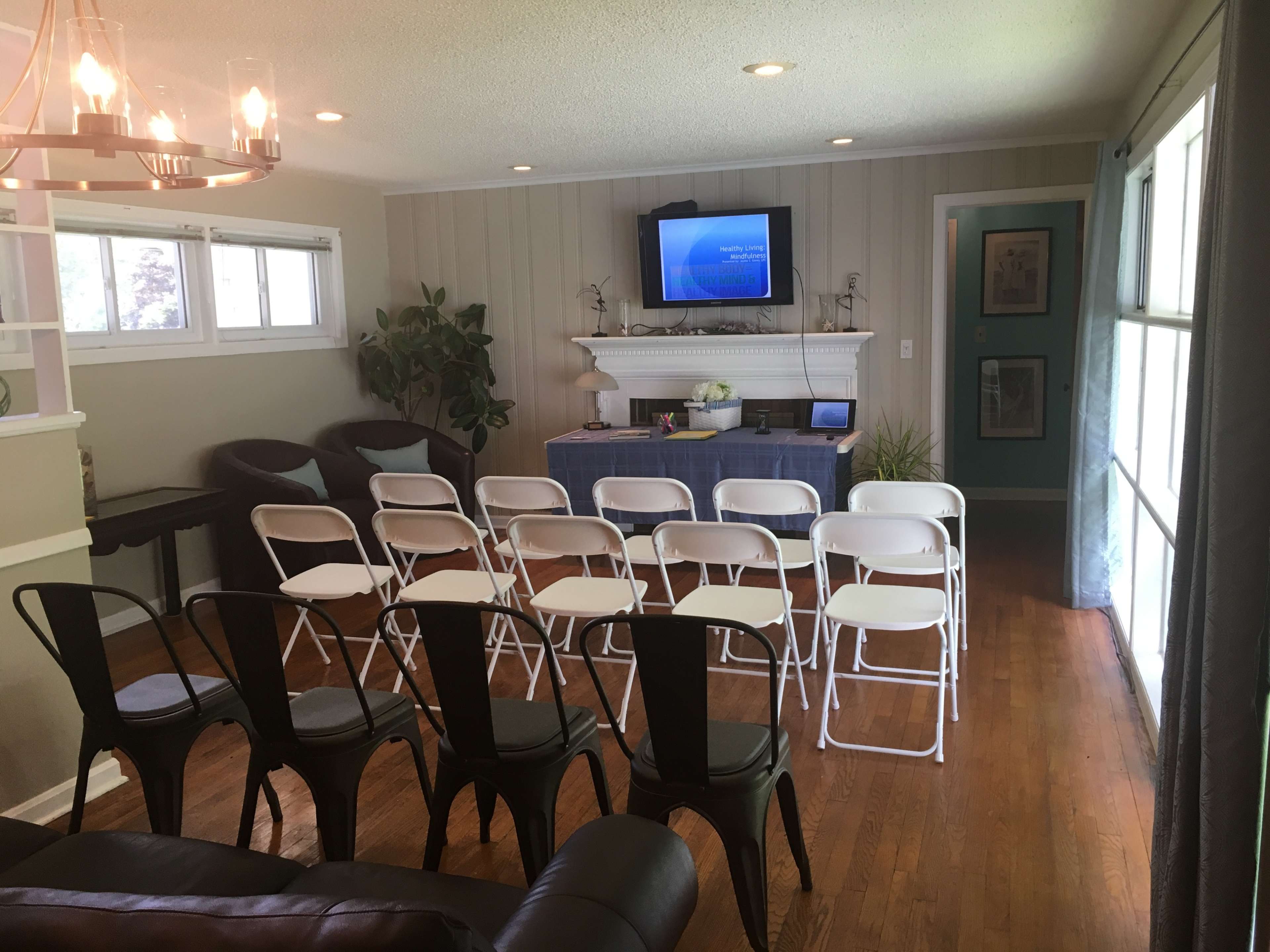 A living room is set up with rows of white folding chairs facing a table and a wall-mounted screen.