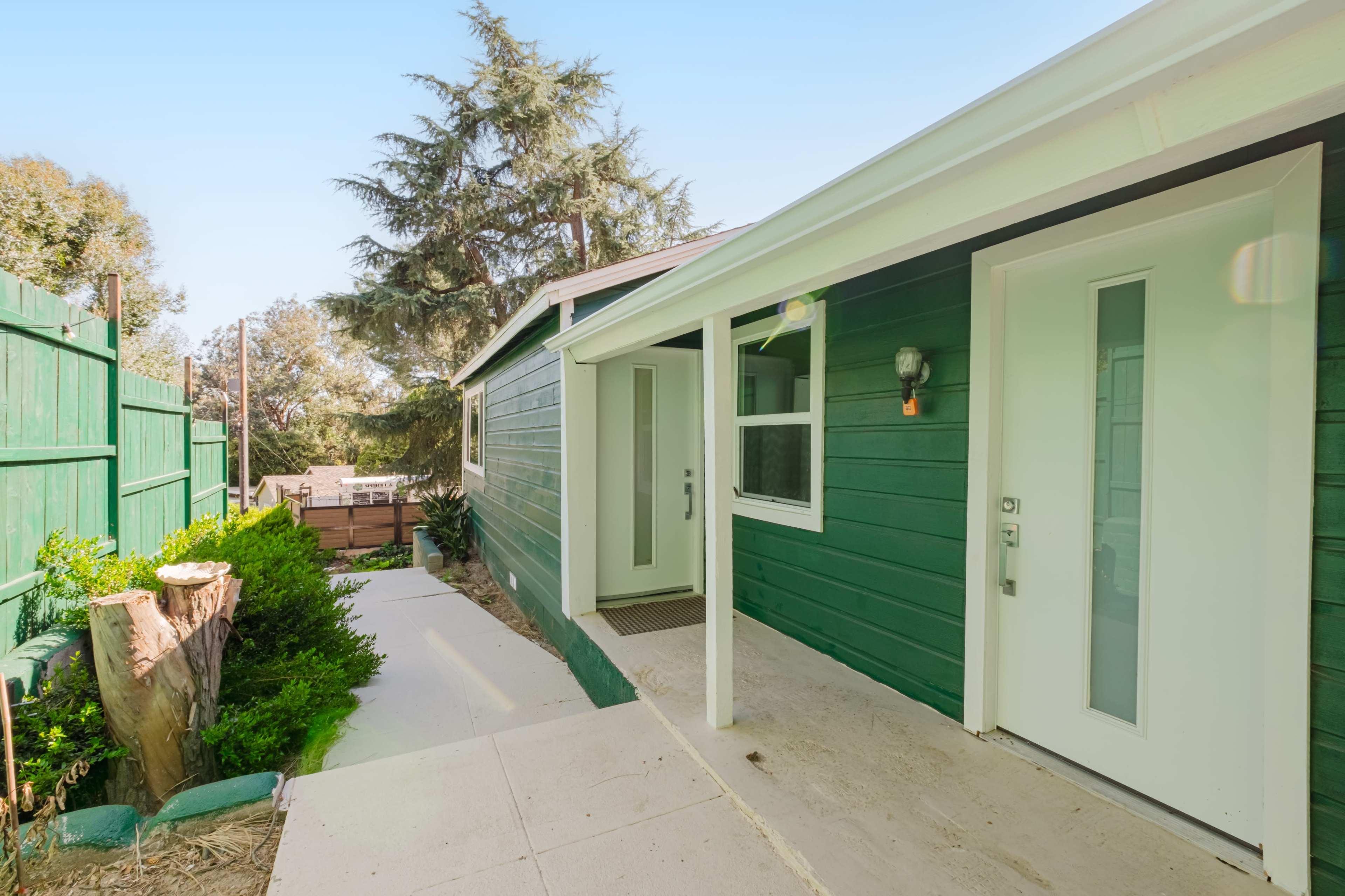 A green wooden house with a concrete walkway leading to a white front door and a corner garden area.