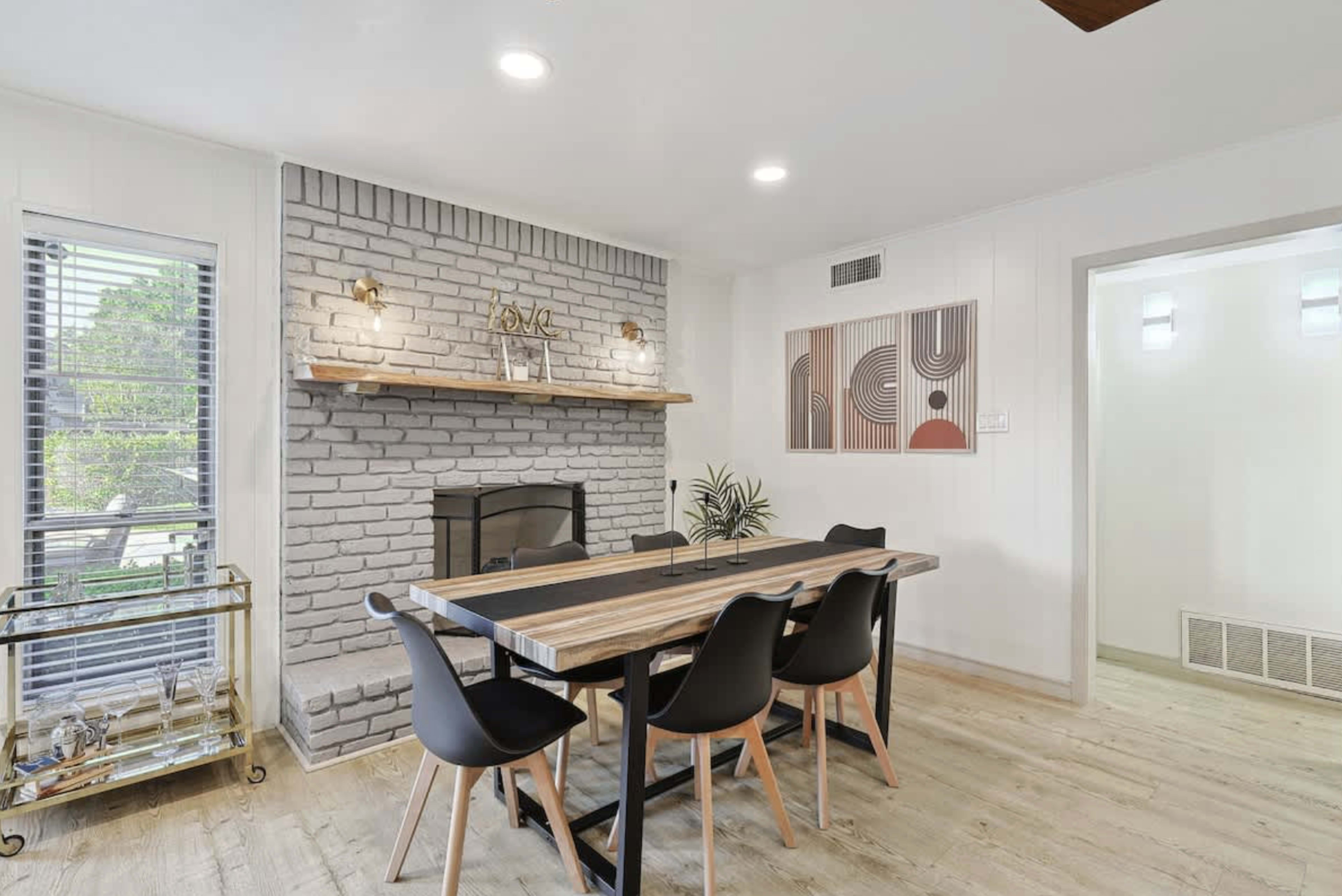 A dining area features a wooden table surrounded by black chairs, a gray brick fireplace, and a decorative wall art piece.