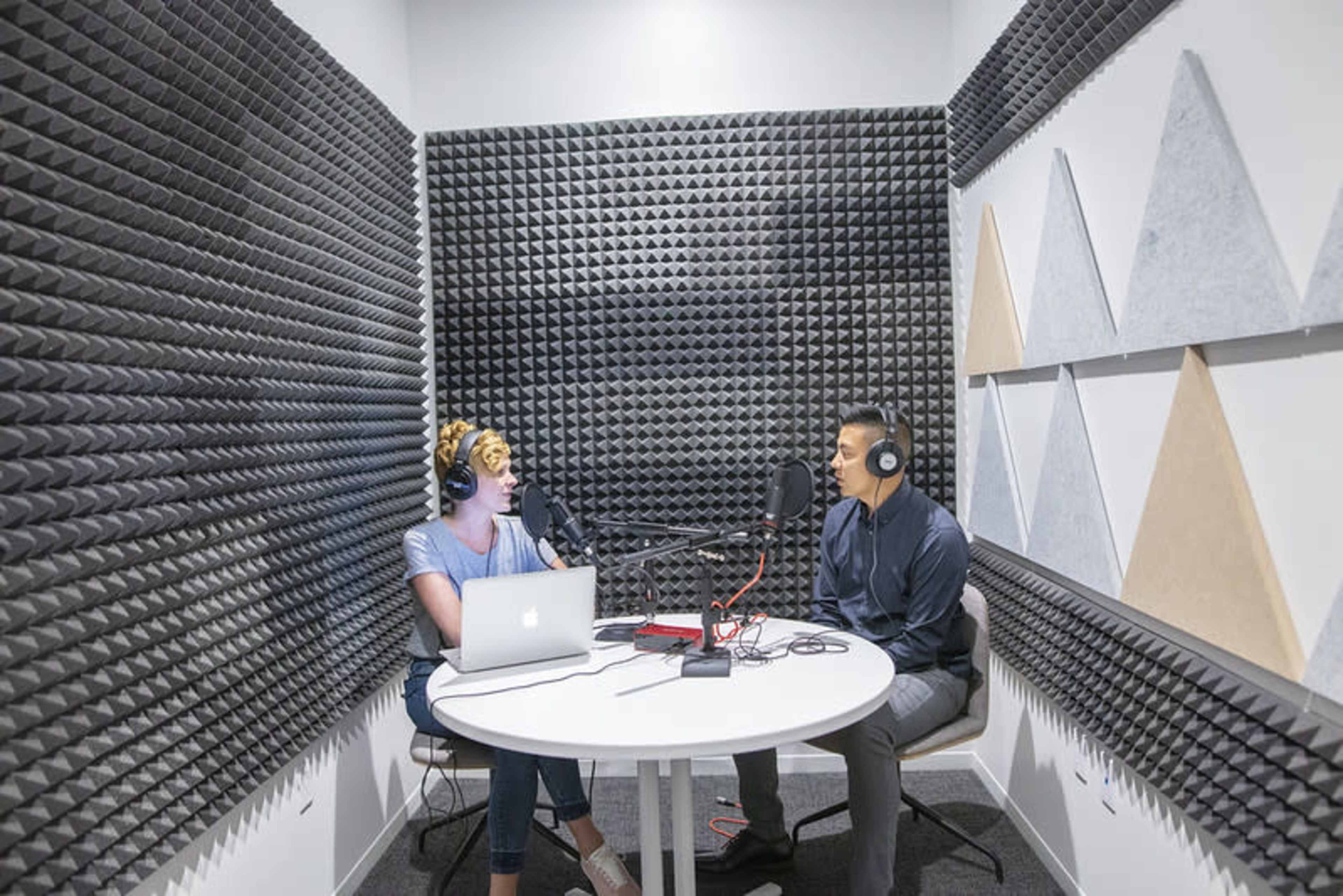 Two individuals are seated at a round table in a soundproof recording studio, engaging in a conversation while using microphones and headphones.
