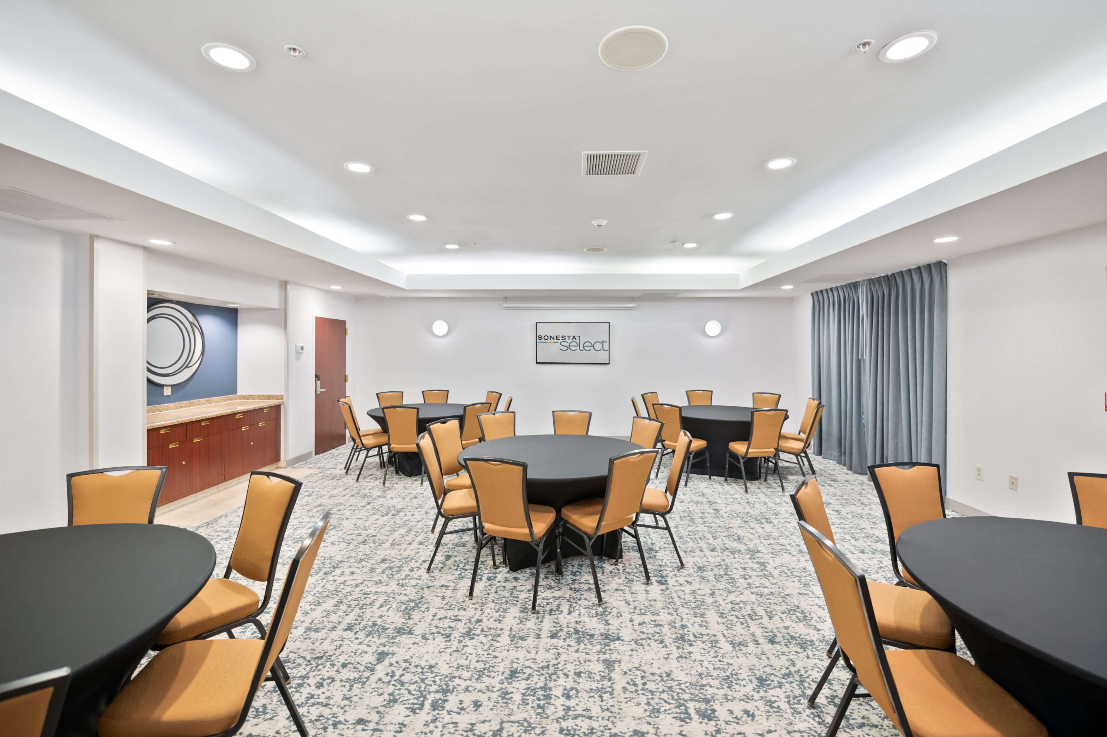 The image shows a conference room set up with multiple round tables covered in black tablecloths and surrounded by orange chairs, along with a kitchenette area in the background.