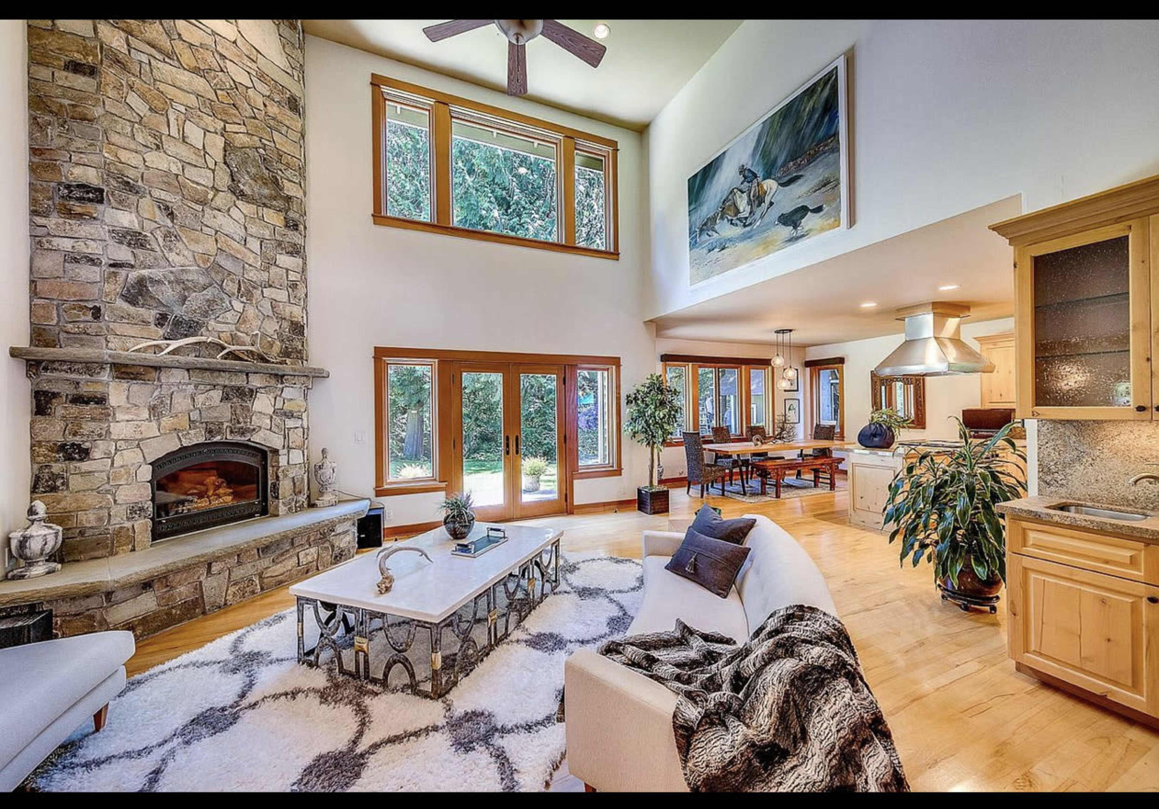 The image shows a spacious living room featuring a large stone fireplace, hardwood flooring, a white sofa, and large windows allowing natural light to enter.