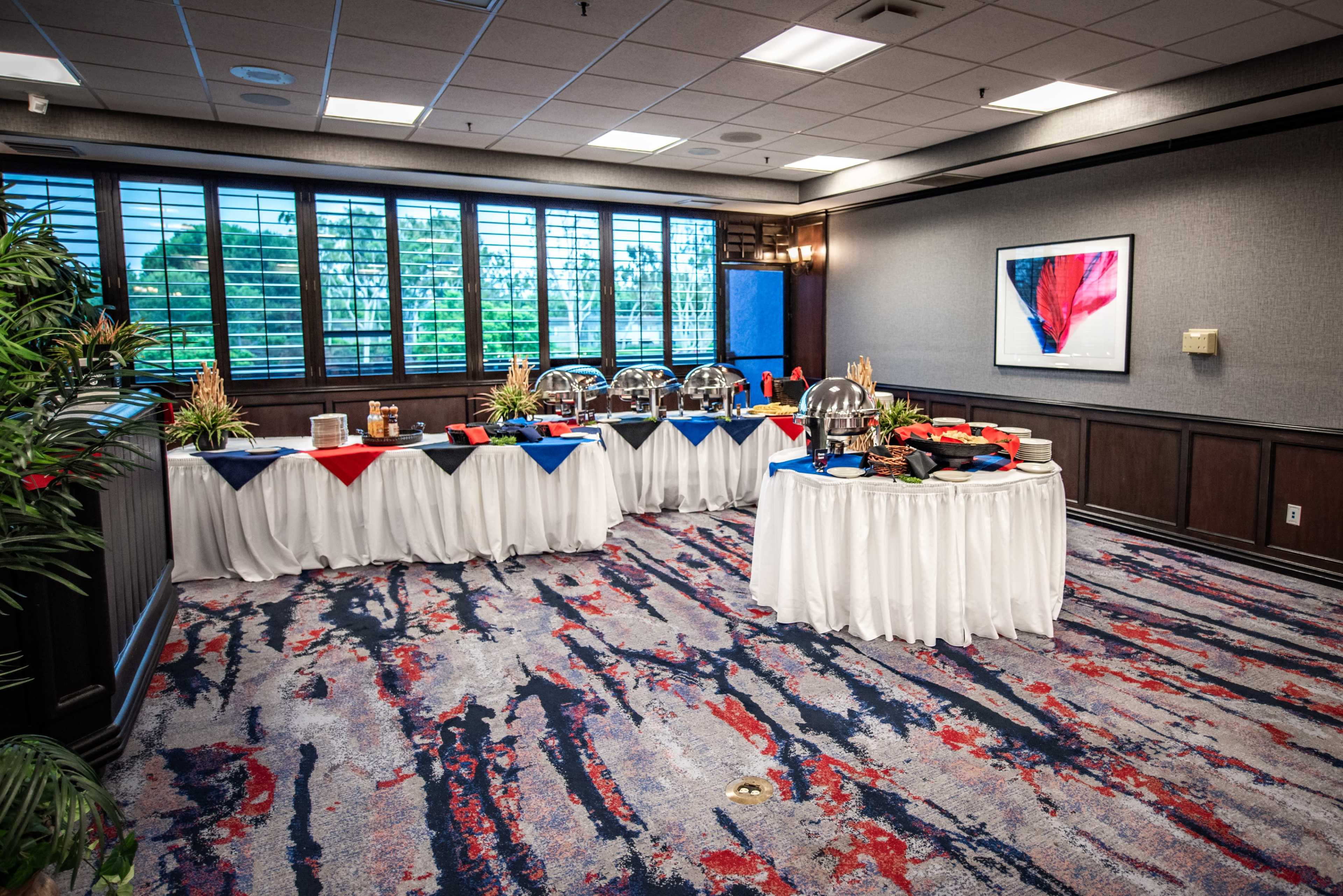 A banquet room features a buffet setup with various food items displayed on two tables covered with white tablecloths, surrounded by a red and blue color scheme.