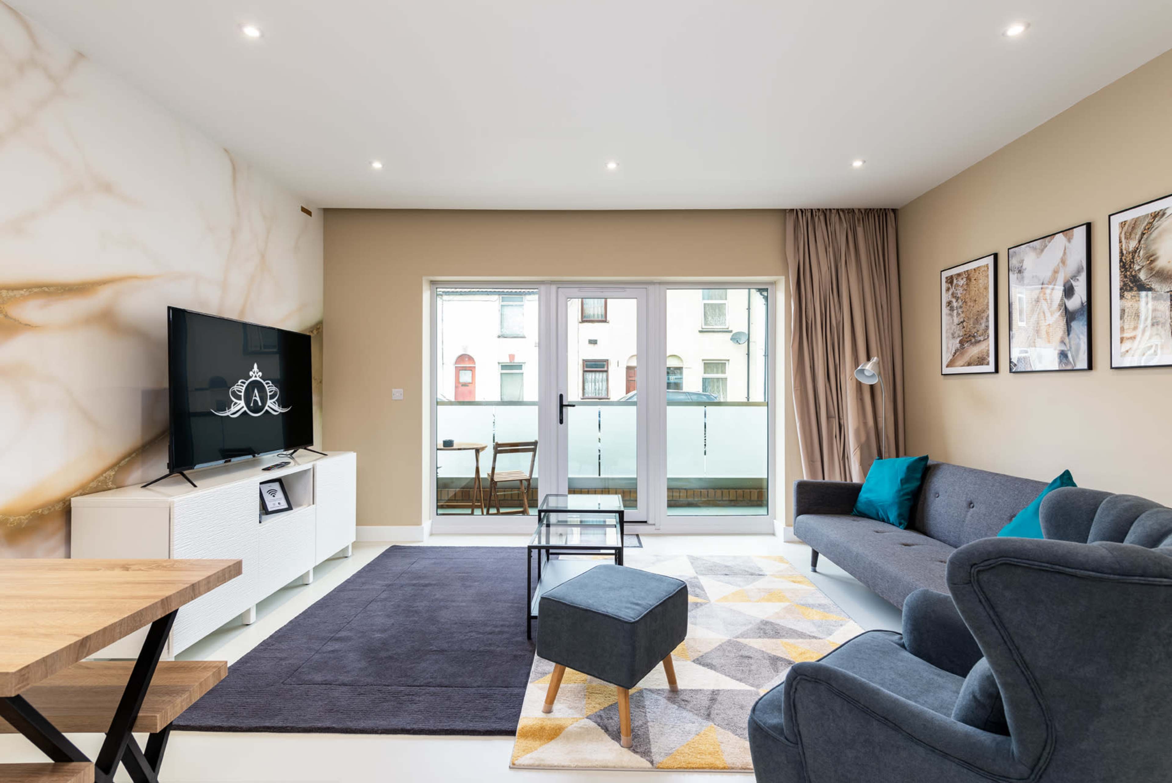 A modern living room features a large TV mounted on a marble-patterned wall, a gray sofa, a glass coffee table, and a balcony with outdoor seating visible through sliding doors.