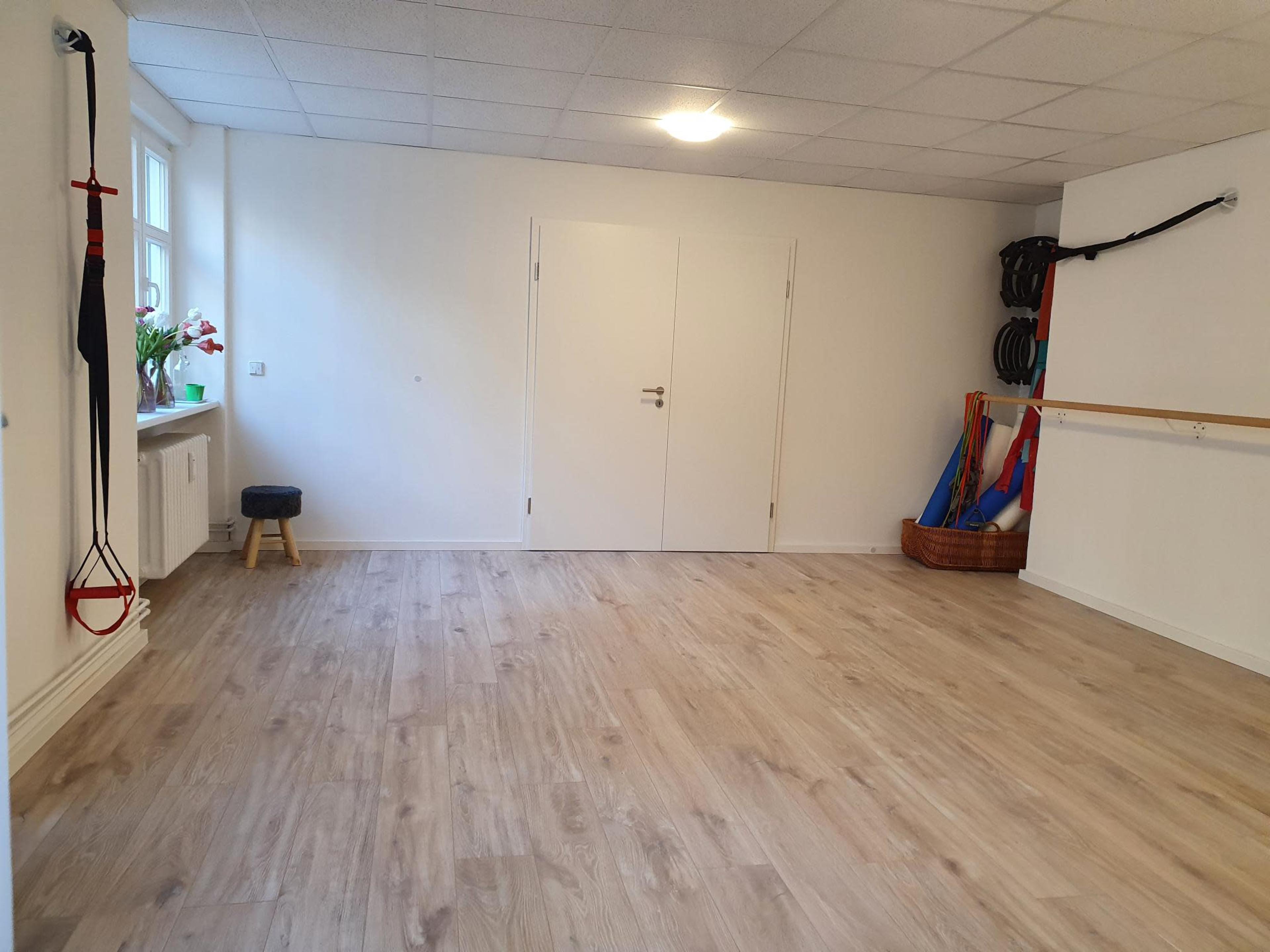 The image shows a spacious, empty room with wooden flooring, a large mirror, and exercise equipment against the walls.