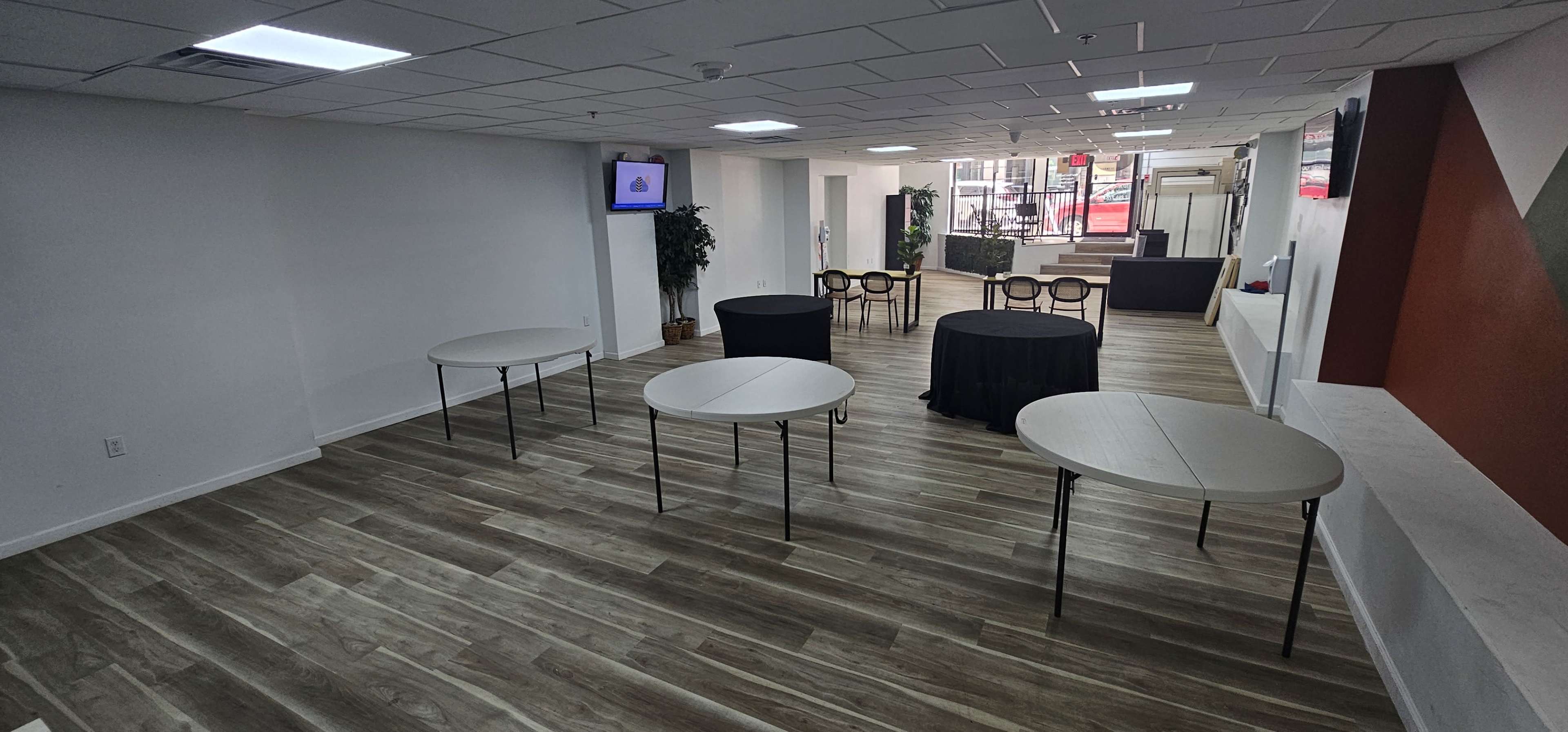 The image shows an empty room with wooden flooring, featuring several round and rectangular tables arranged in a setup for an event, along with a screen mounted on the wall and a small seating area in the background.