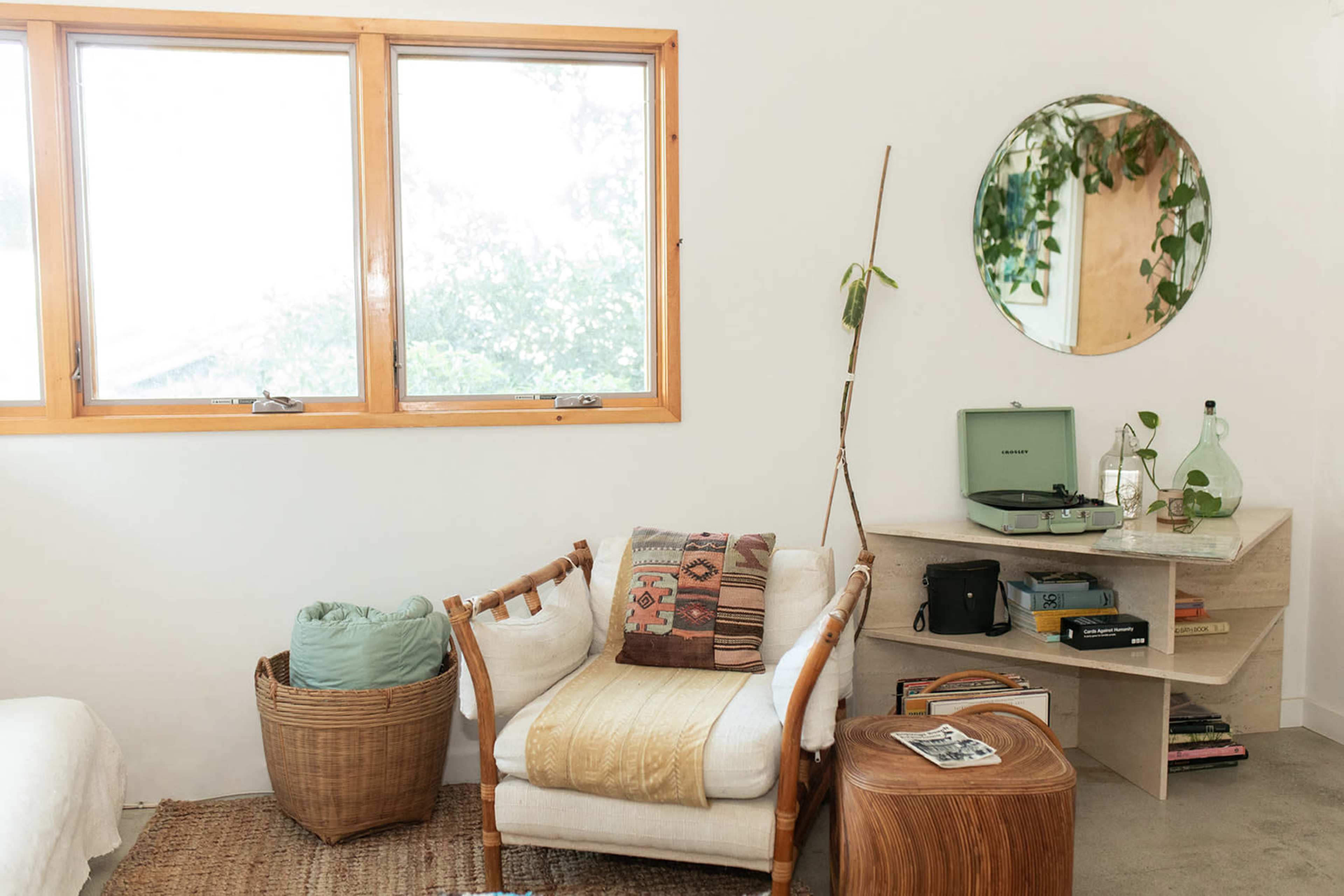 The image shows a cozy living space featuring a chair with a patterned throw, a small wooden side table, a record player on a shelf, and large windows that let in natural light.
