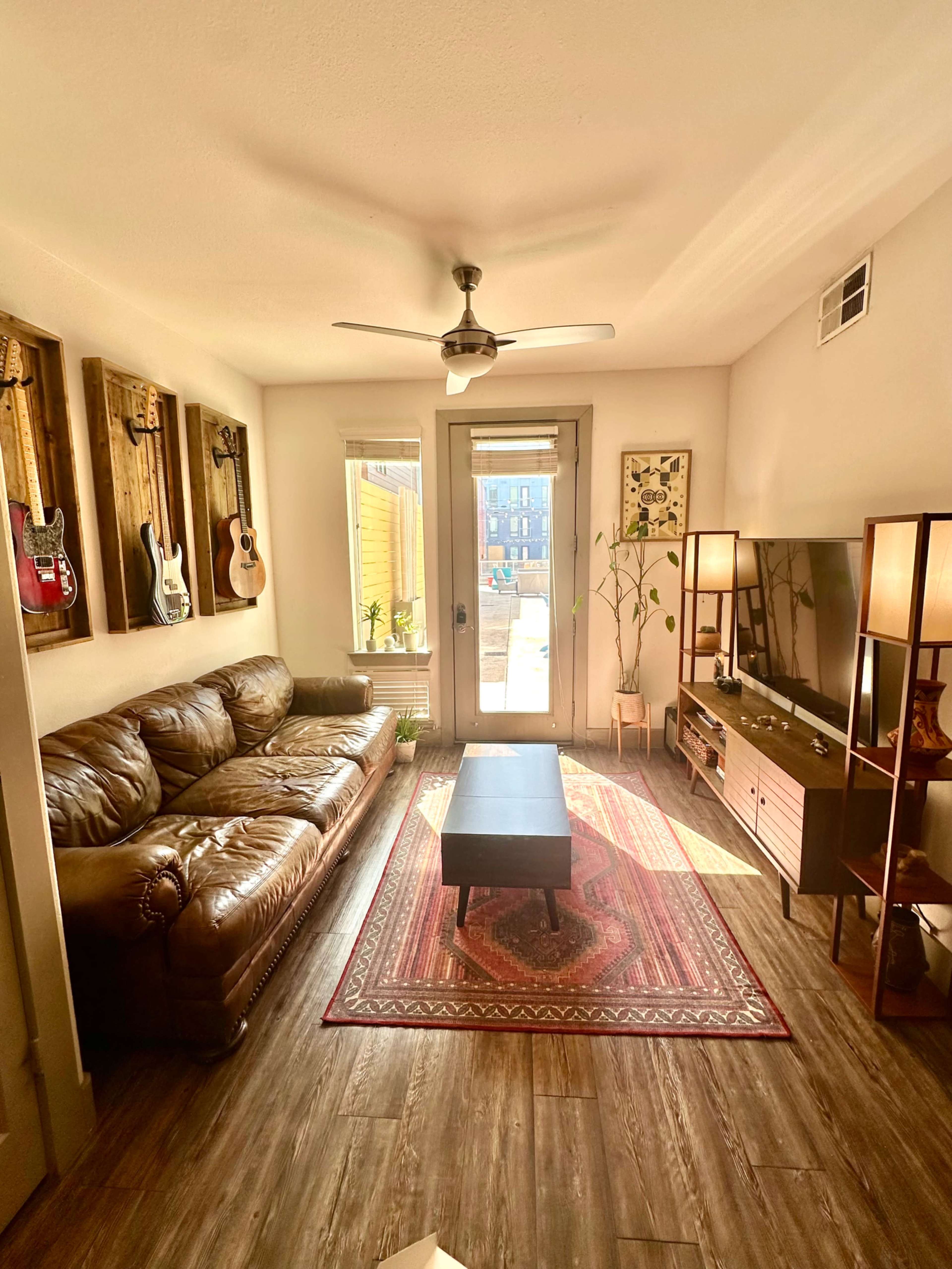 The living room features a brown leather sofa, a coffee table, a television stand with a TV, and decorative guitars on the wall, illuminated by natural light from a door leading outside.
