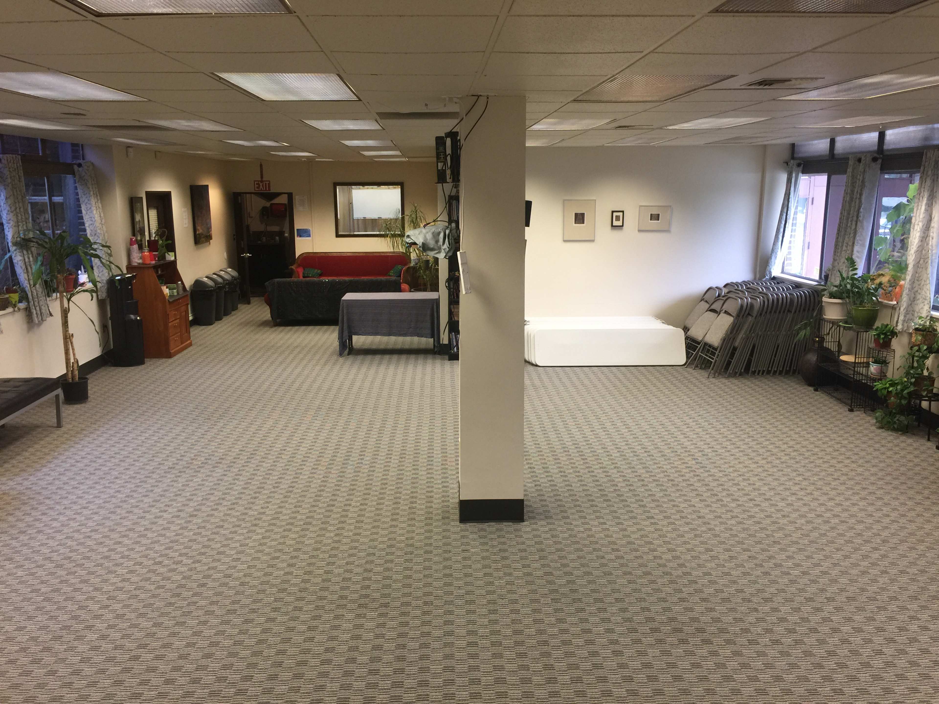 A spacious, empty room with carpeted flooring, featuring a few pieces of furniture, plants, and folding chairs stacked against the wall.