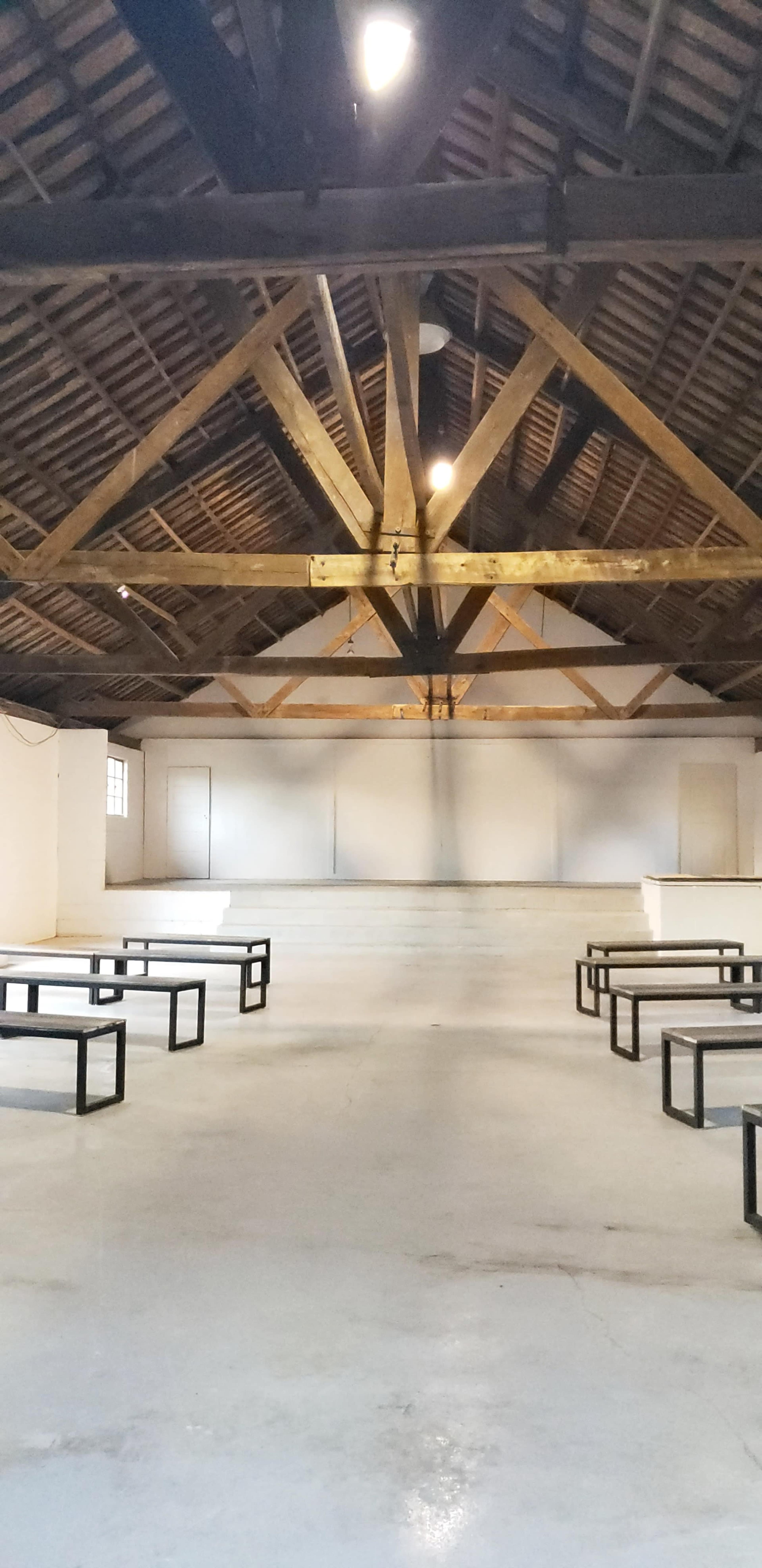 An empty hall with a wooden beamed ceiling and several black metal benches arranged on a polished concrete floor.