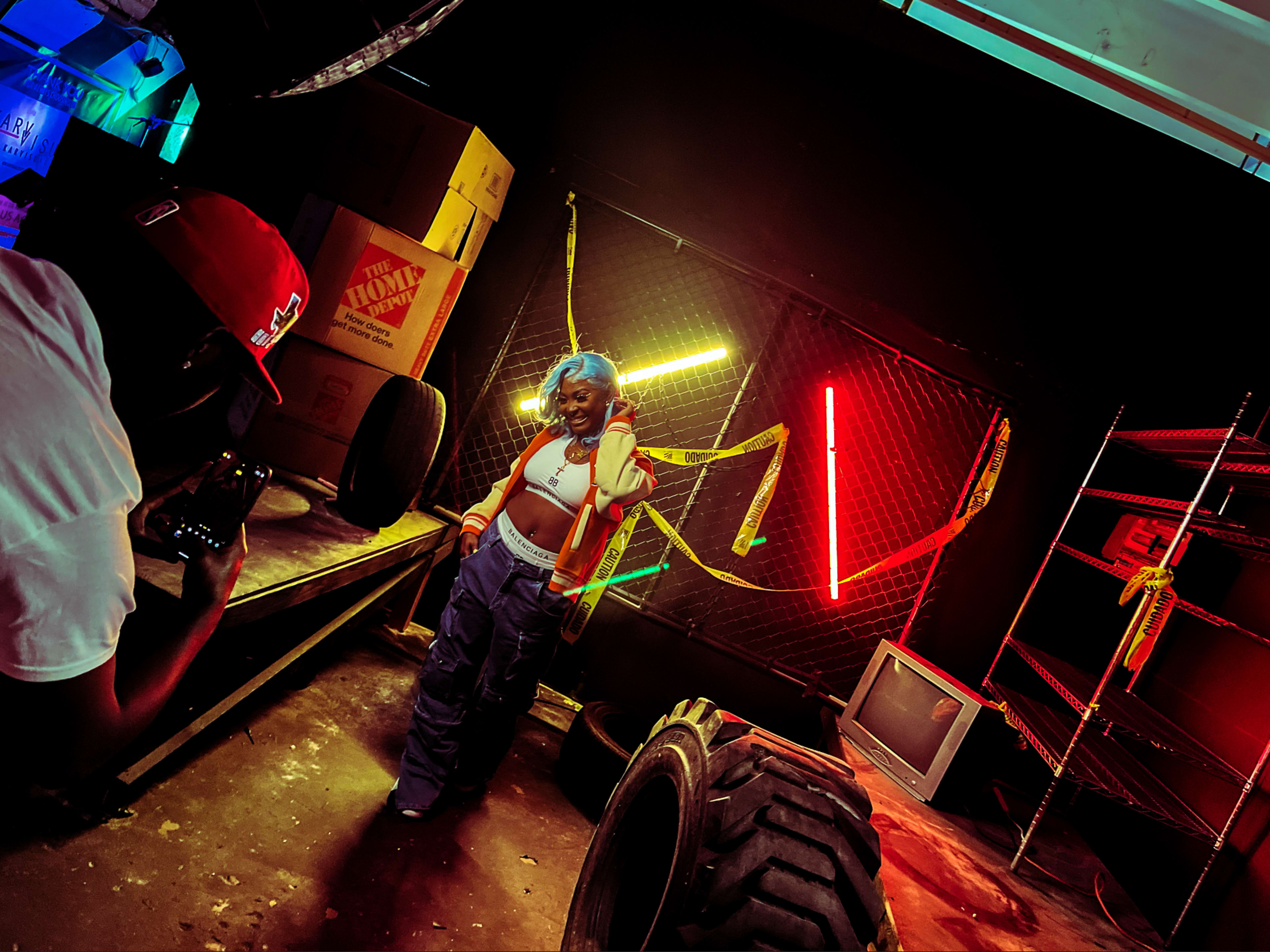 A person poses in front of neon lights while another individual takes a photograph in a dimly lit space filled with boxes and tires.