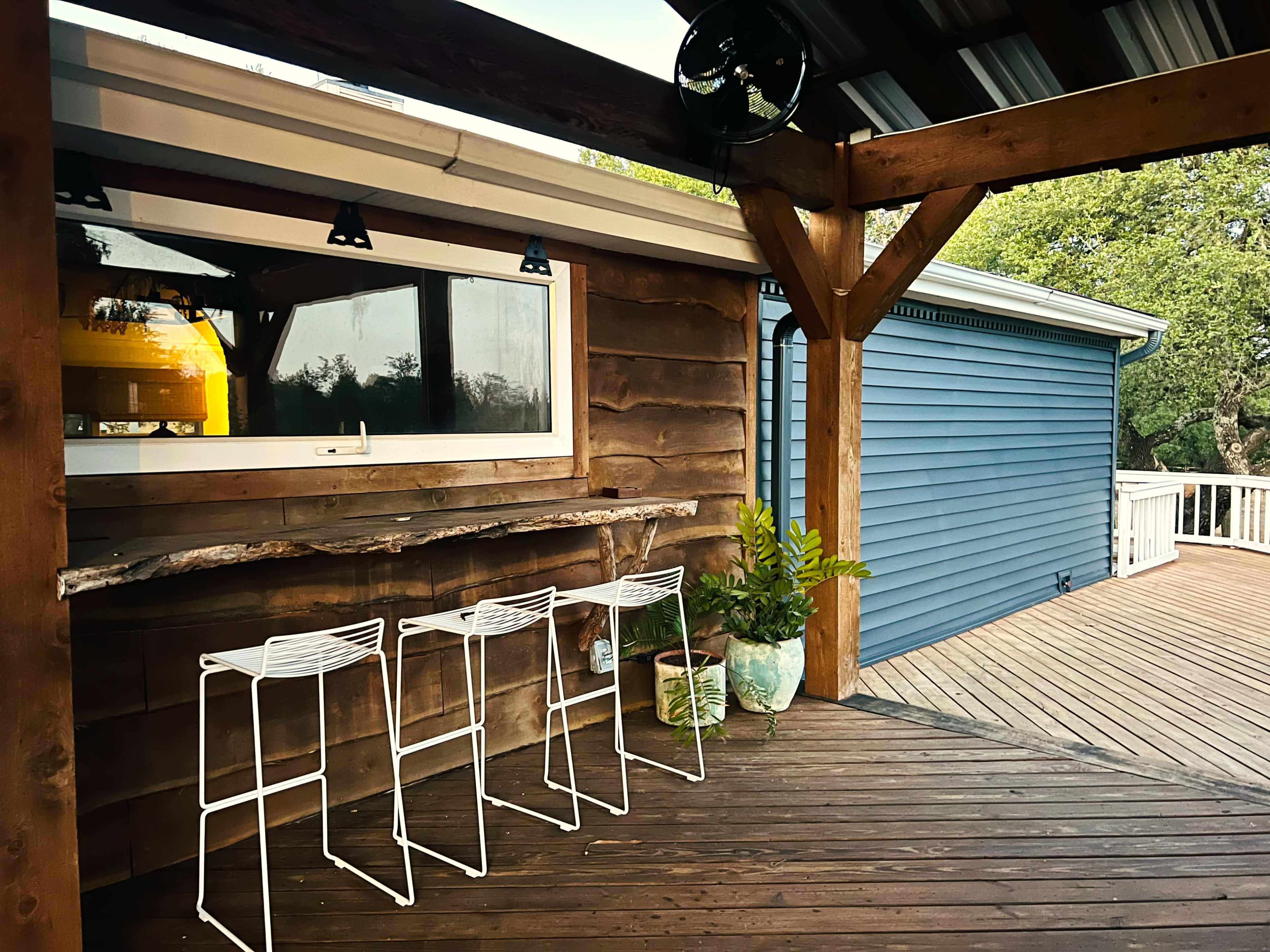 The image shows a wooden deck with three white metal bar stools positioned in front of a rustic wooden counter, beside a blue-painted wall of a house.