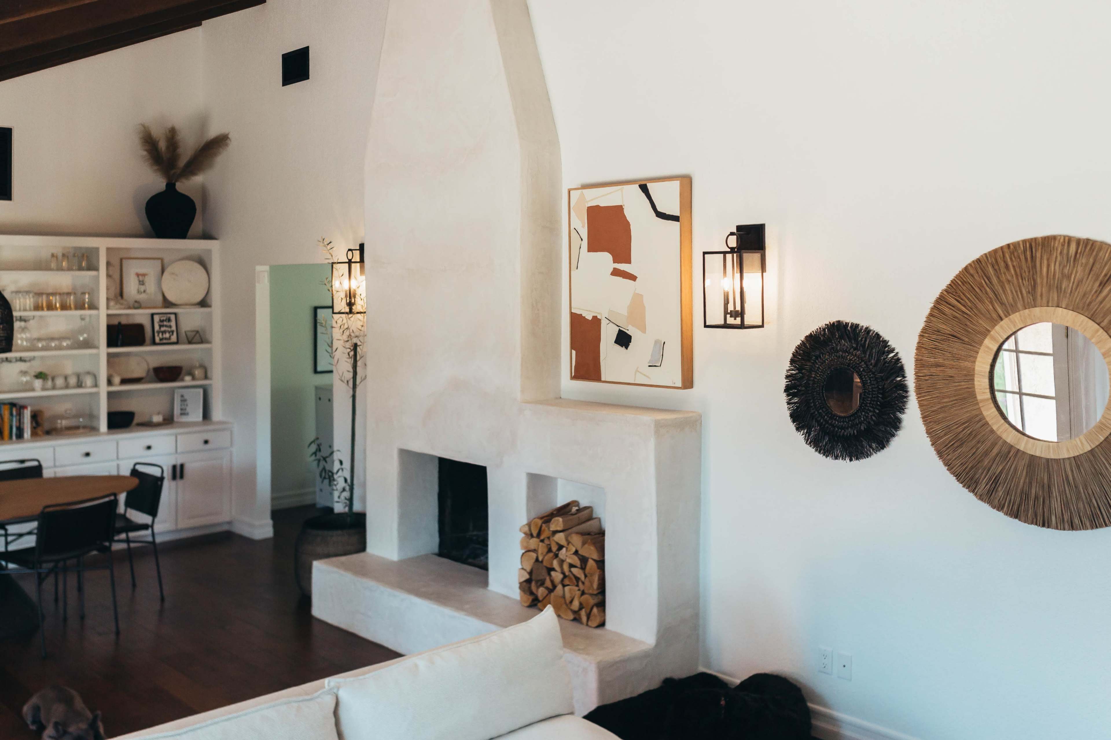 A modern living room featuring a white plaster fireplace, a piece of artwork on the wall, and decorative mirrors beside a stacked pile of firewood.