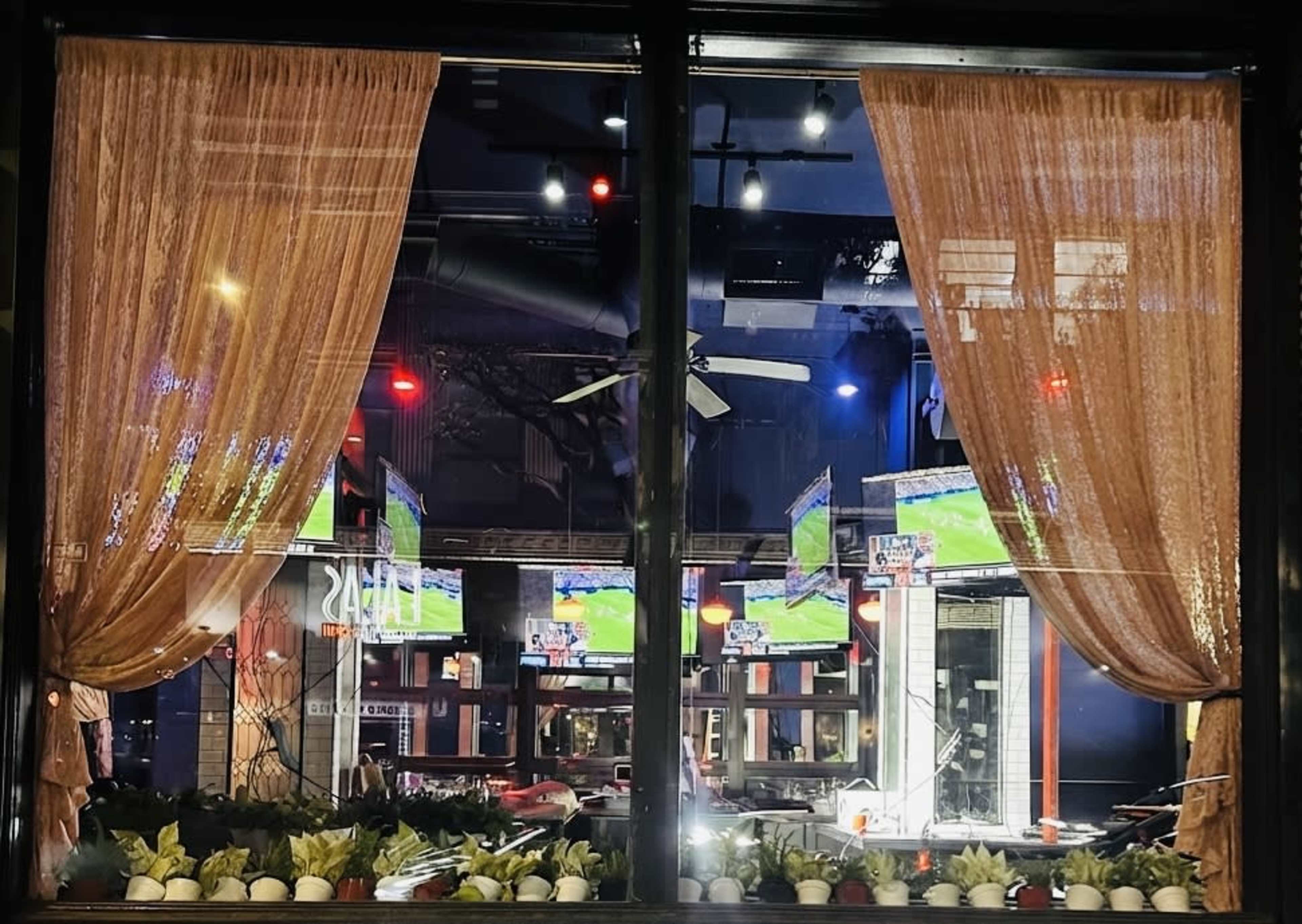 The image shows a restaurant window with sheer curtains, displaying multiple television screens showing a sports game and illuminated by various colored lights.